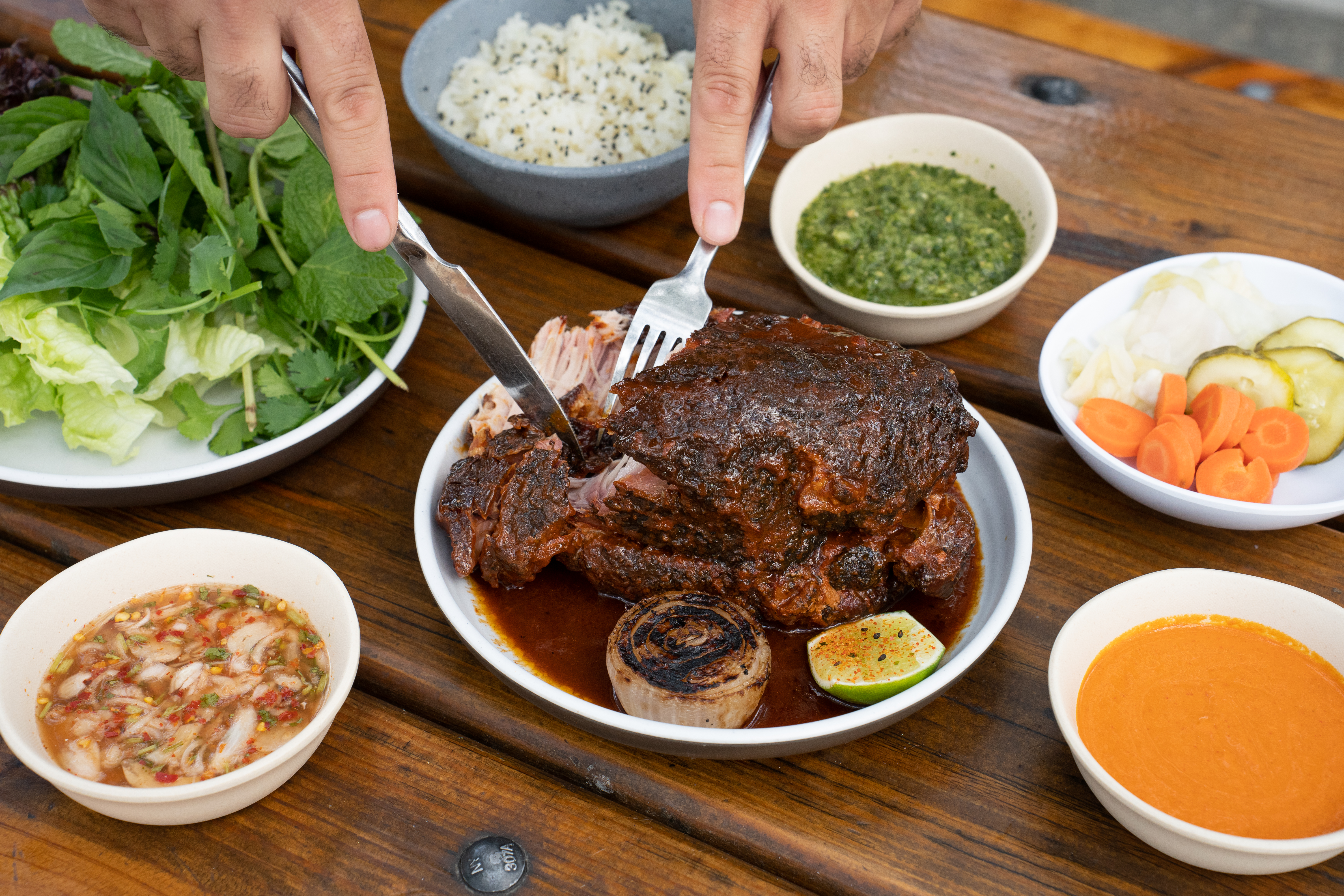 A pair of hands hold a fork and knife and cut into a pork butt that’s covered in marinade. Numerous traditional Asian sides in small bowls surround the plate.