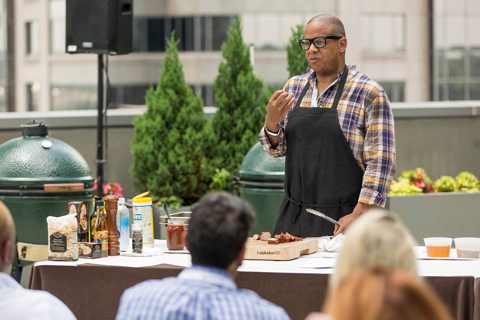Todd Richards speaking to a crowd outside on the terrace of the Lowes Hotel in Midtown Atlanta during a 2018 Food and Wine Festival session. Richards is wearing stylish thick, black rimmed glasses, a plaid button down shirt, and a black apron tied in the front