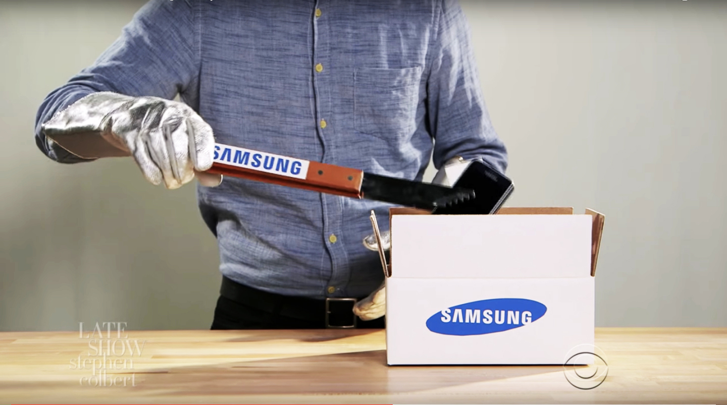 A clip from Colbert's video in which you see a man wearing aluminum gloves, holding tongs emblazoned with the Samsung logo, gently putting a phone into a Samsung box. 