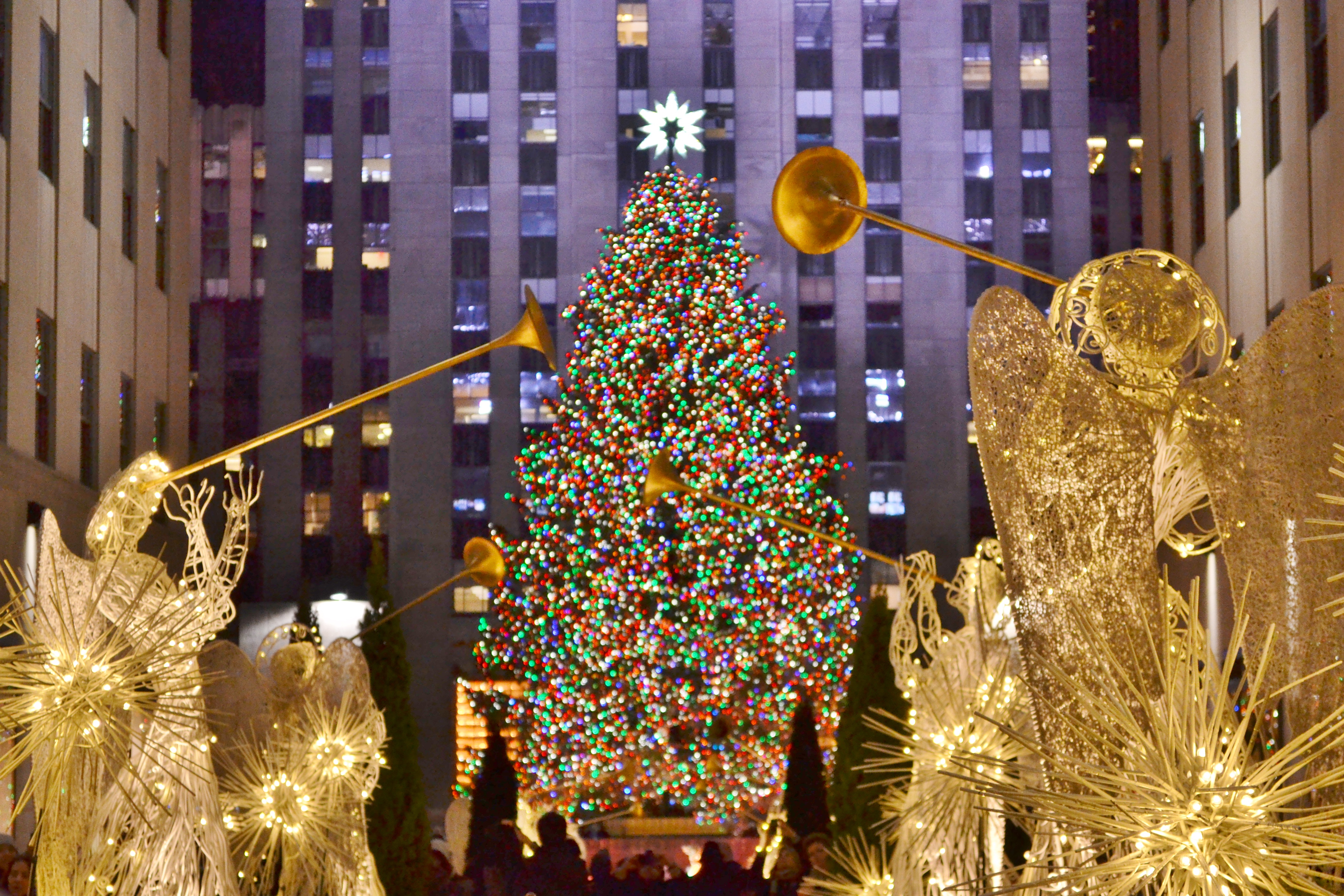 The Rockefeller Center Christmas tree. The tree is decorated with lights. There are illuminated statues of angels with trumpets in front of the tree.