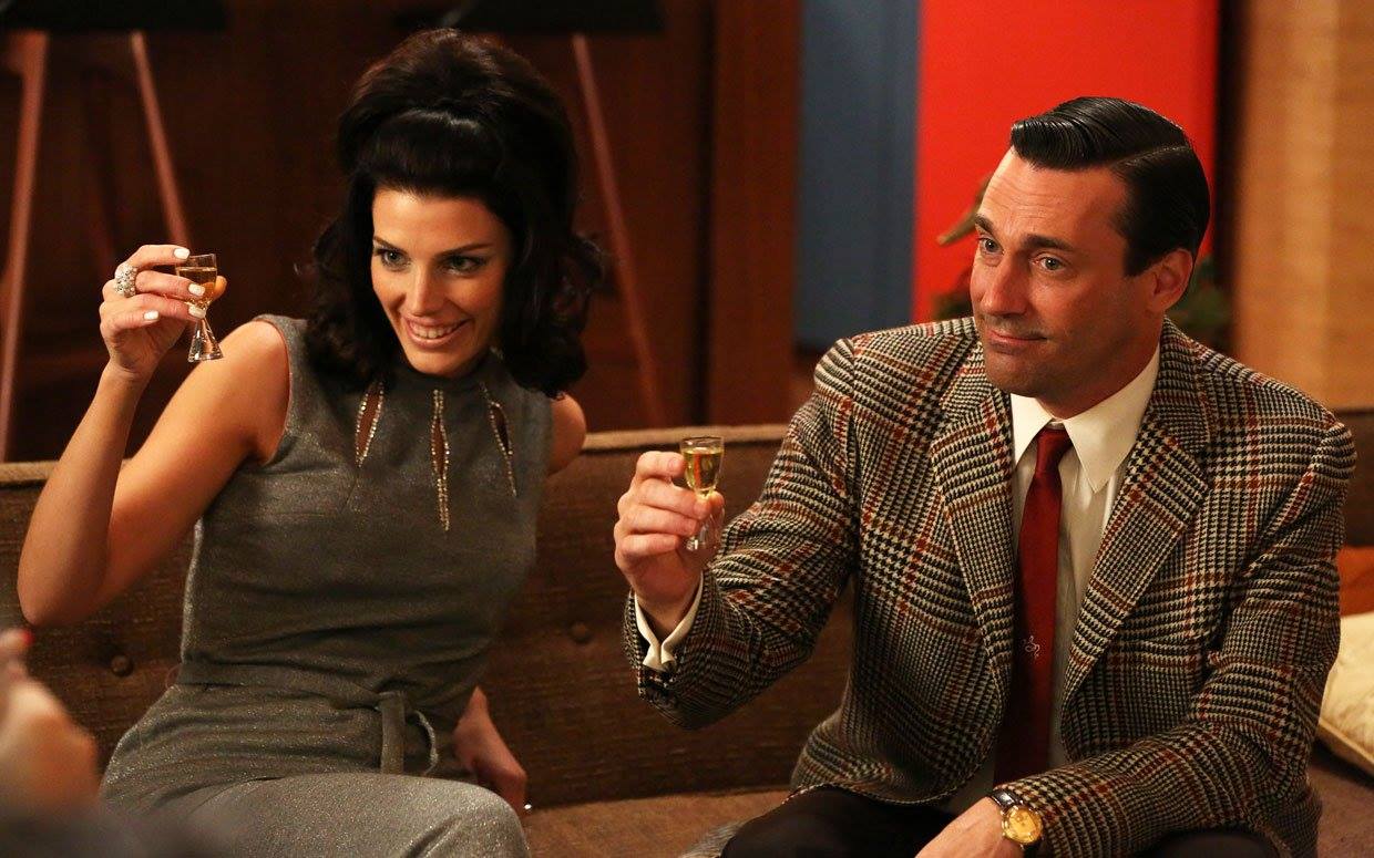 Characters Megan and Don Draper from TV show Mad Men raising a toast. She has on a sleeveless jumpsuit and he has on a plaid sports coat and tie. They are seated. Her hair is in a semi-bouffant and his is slicked back.