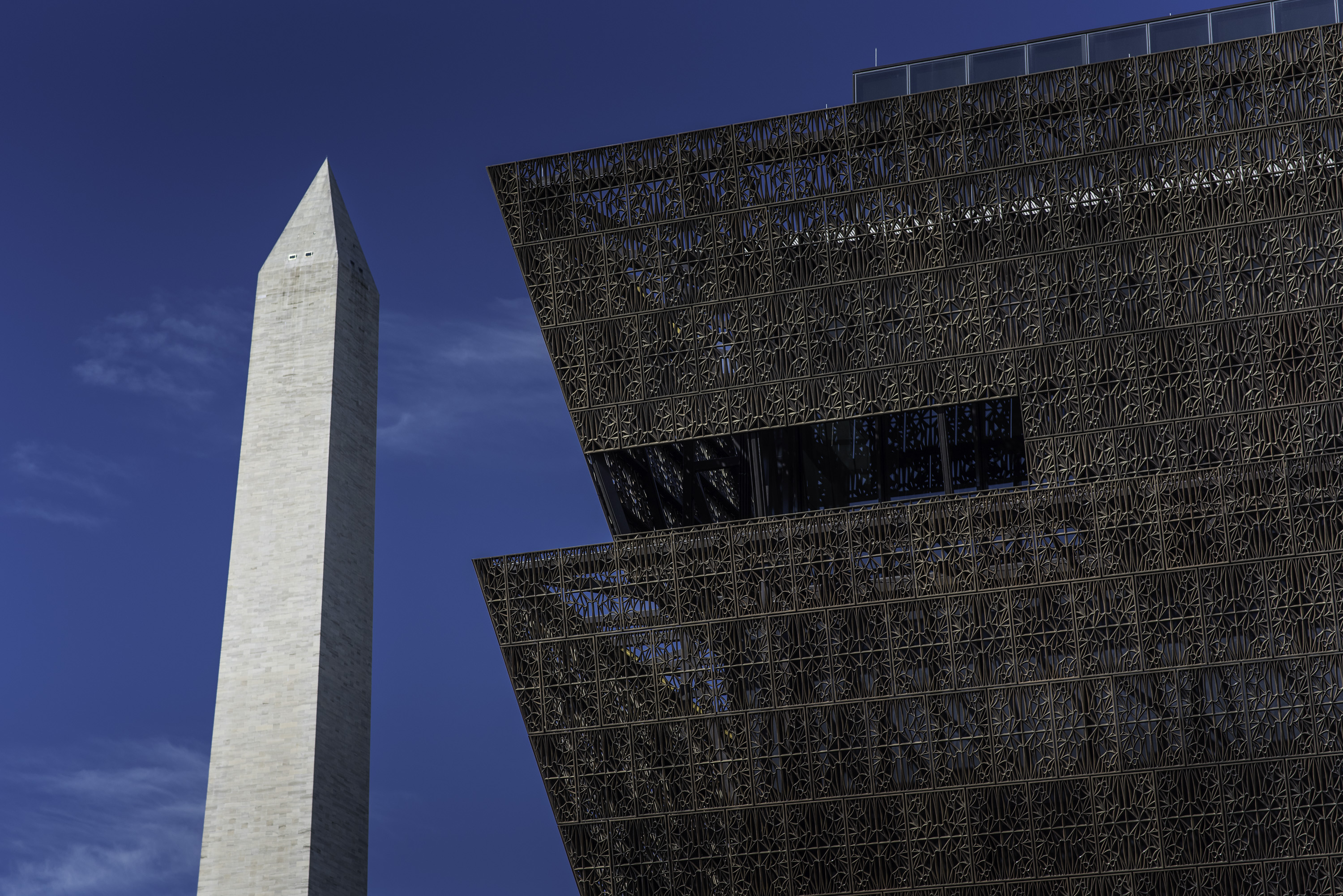 Exterior shot of a corner of a brown steel-framed tiered structure with the obelisk of the Washington Monument in the background. 