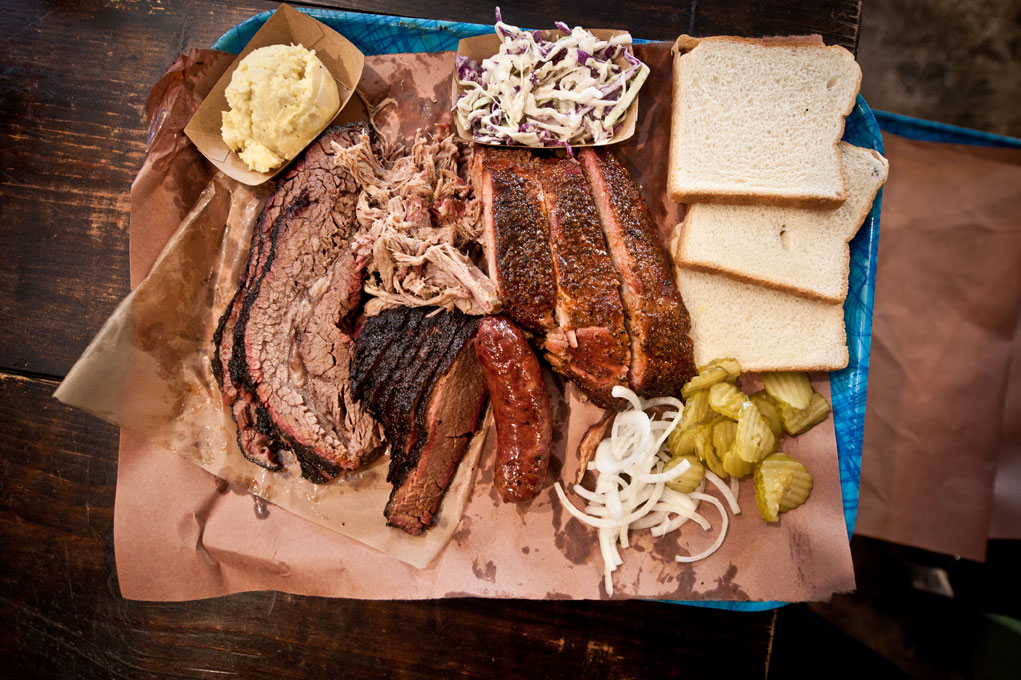 A tray of various barbecue meats ranging from brisket slices to ribs to sausages, alongside sides of coleslaw and potato salad and slices of bread, pickle slices, and onions.