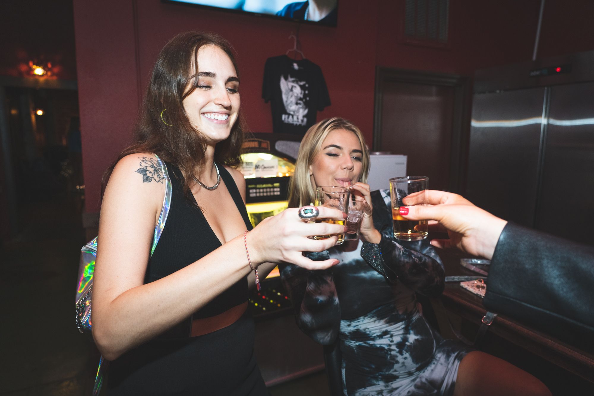 Two women drink shots as a man’s hand holds up a glass to cheers.