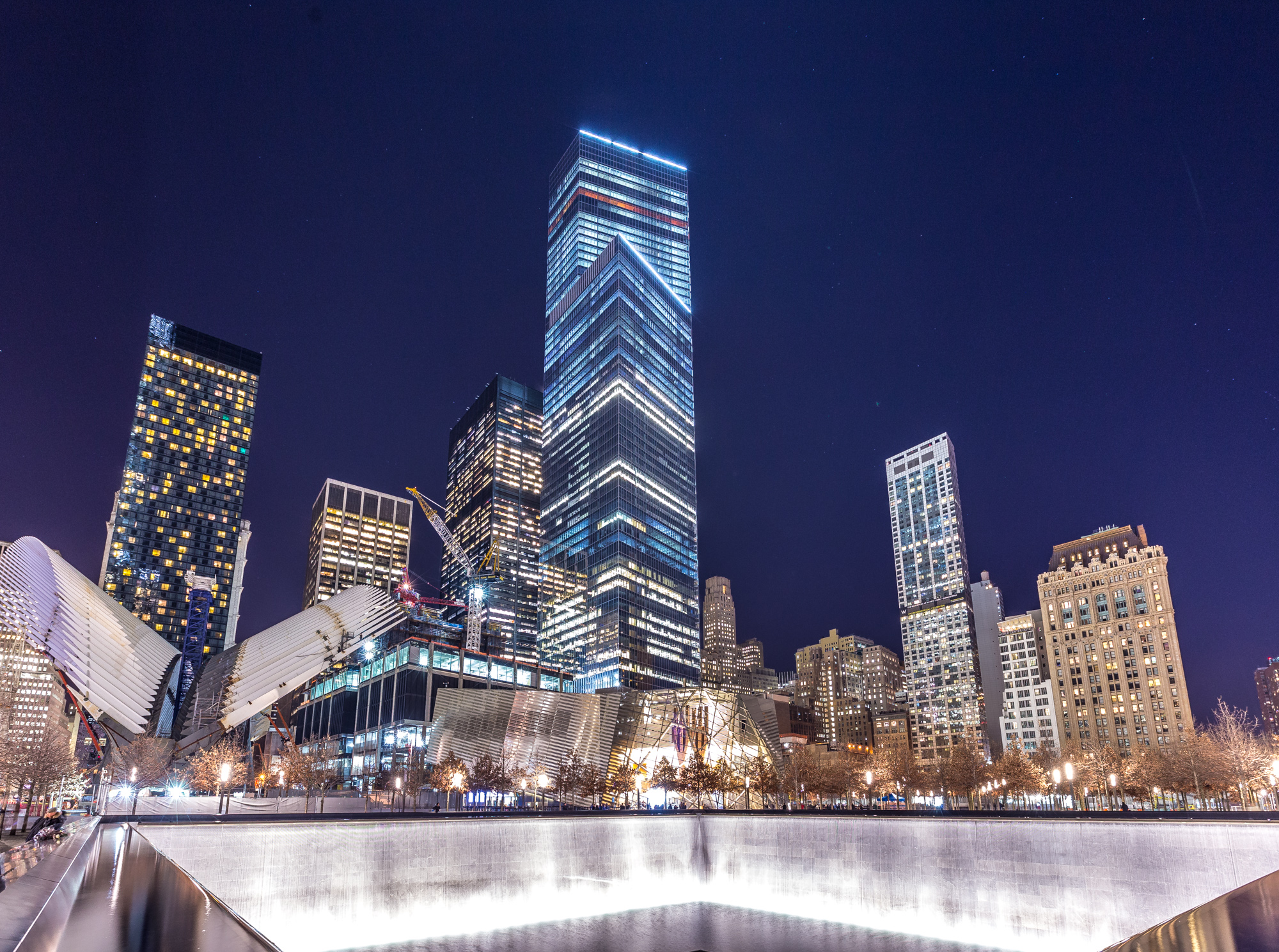 The skyscrapers and buildings at the World Trade Center site. In the foreground is a large memorial fountain.