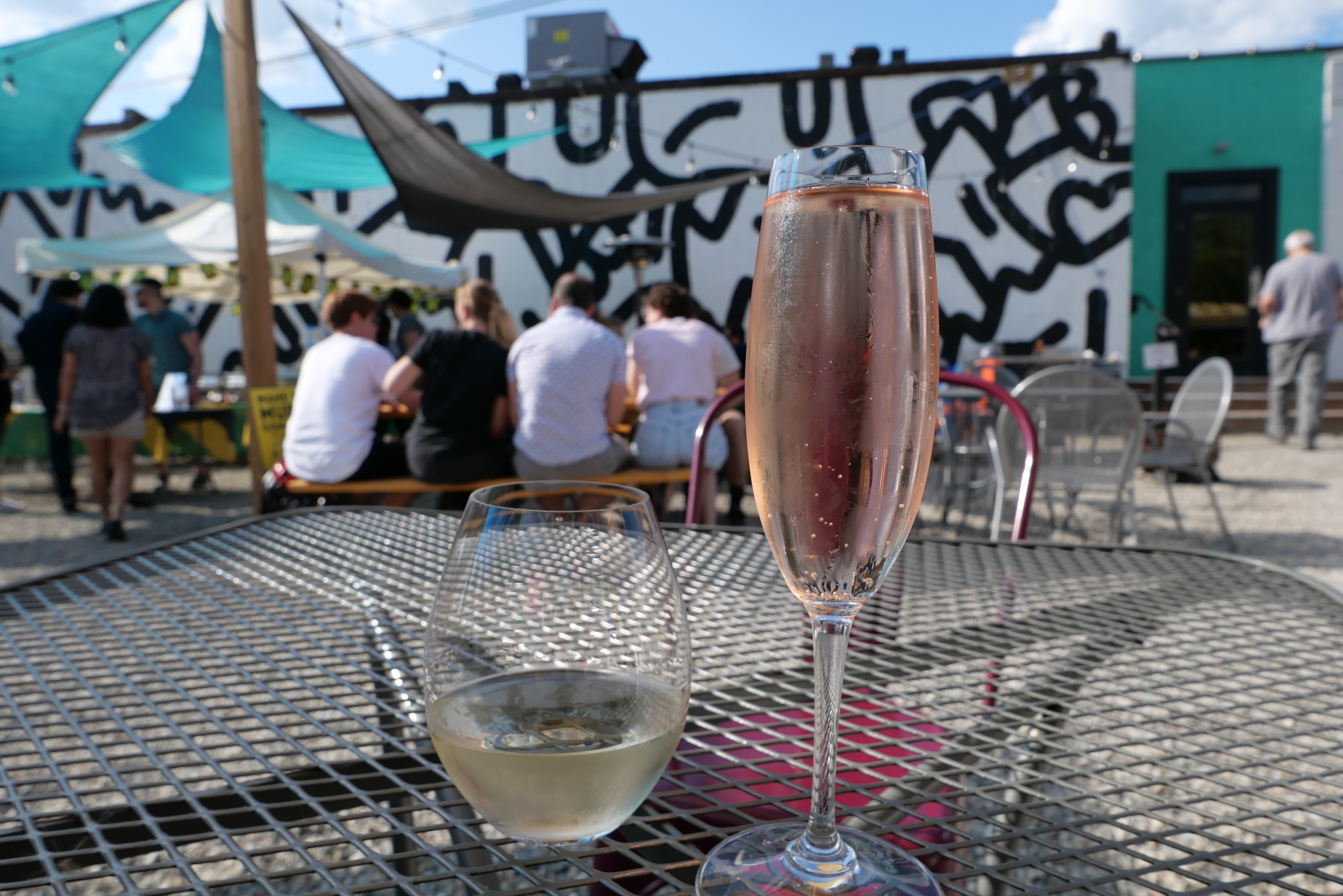 Two glasses of wine, one white and one rose, sit on a metal table outside on a sunlit patio dotted with people congregating at other tables.
