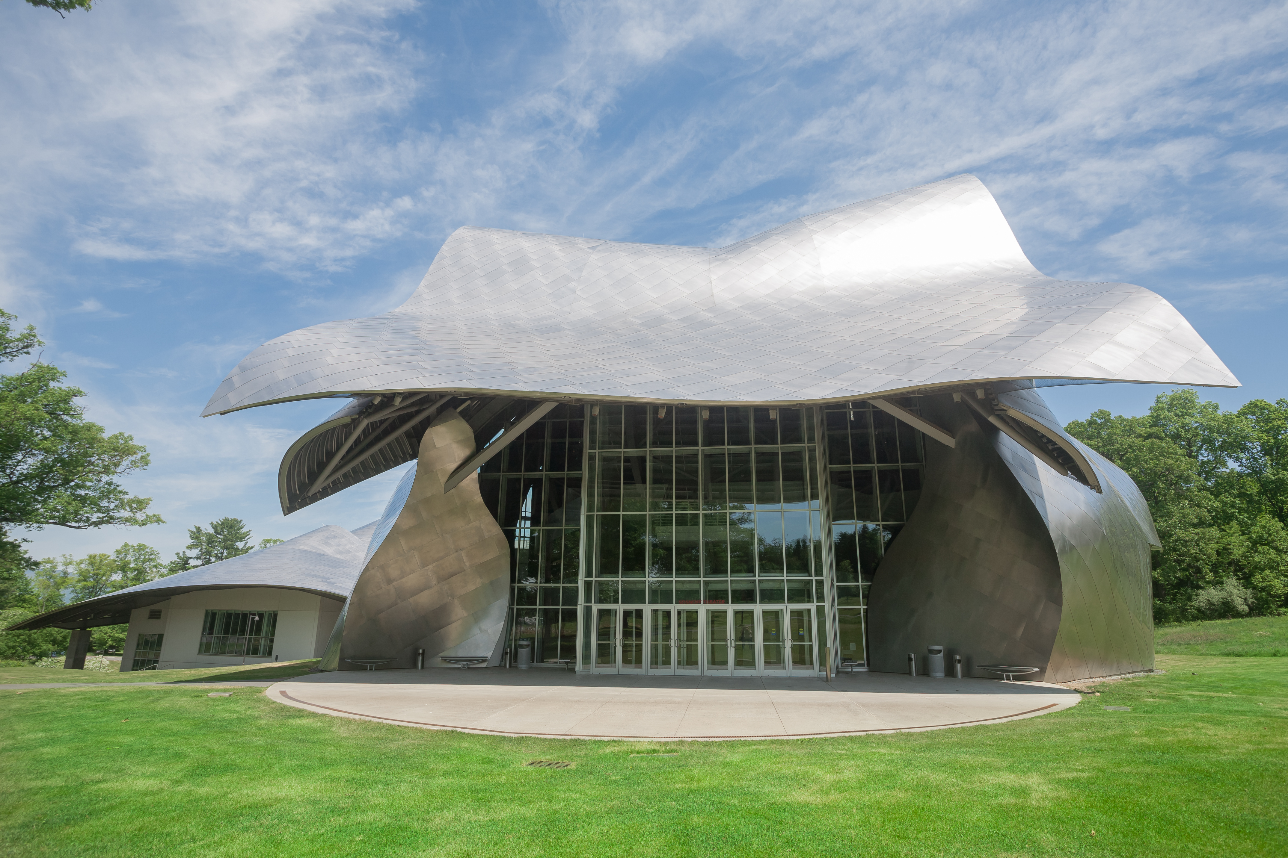The performing arts building at Bard College, the Fisher Center, designed by Frank Gehry. The roof and walls are curved metal. There is a glass entryway. The building is surrounded by a green grass lawn.