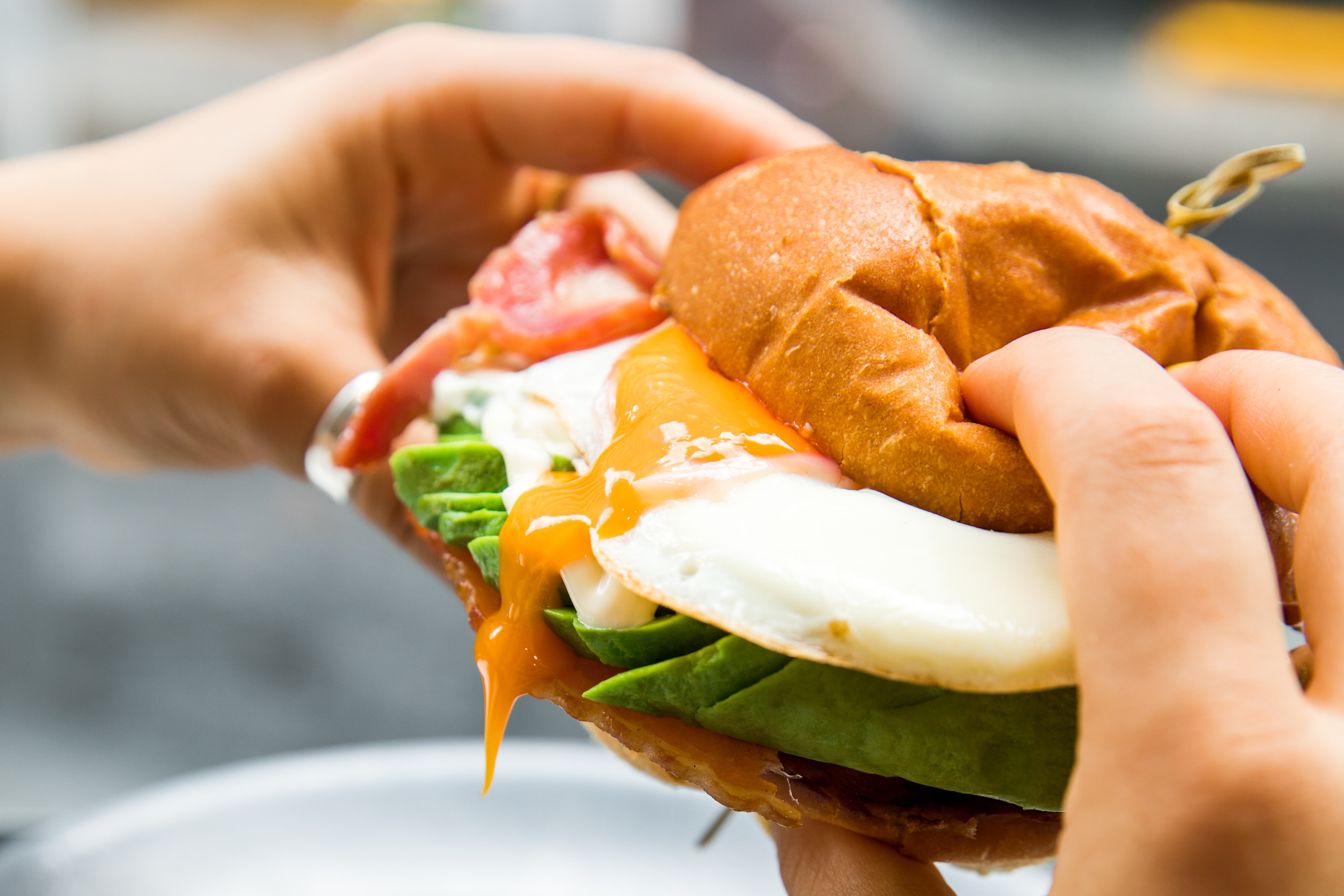 person holding breakfast sandwich with oozing egg and avocado inside.
