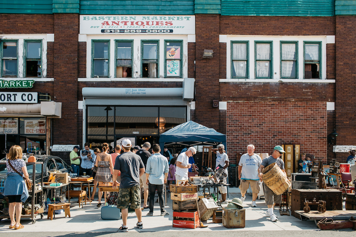 In the foreground are people and an outdoor market in a courtyard. The building in the background is red brick and has a sign on it that reads: Eastern Market antiques.