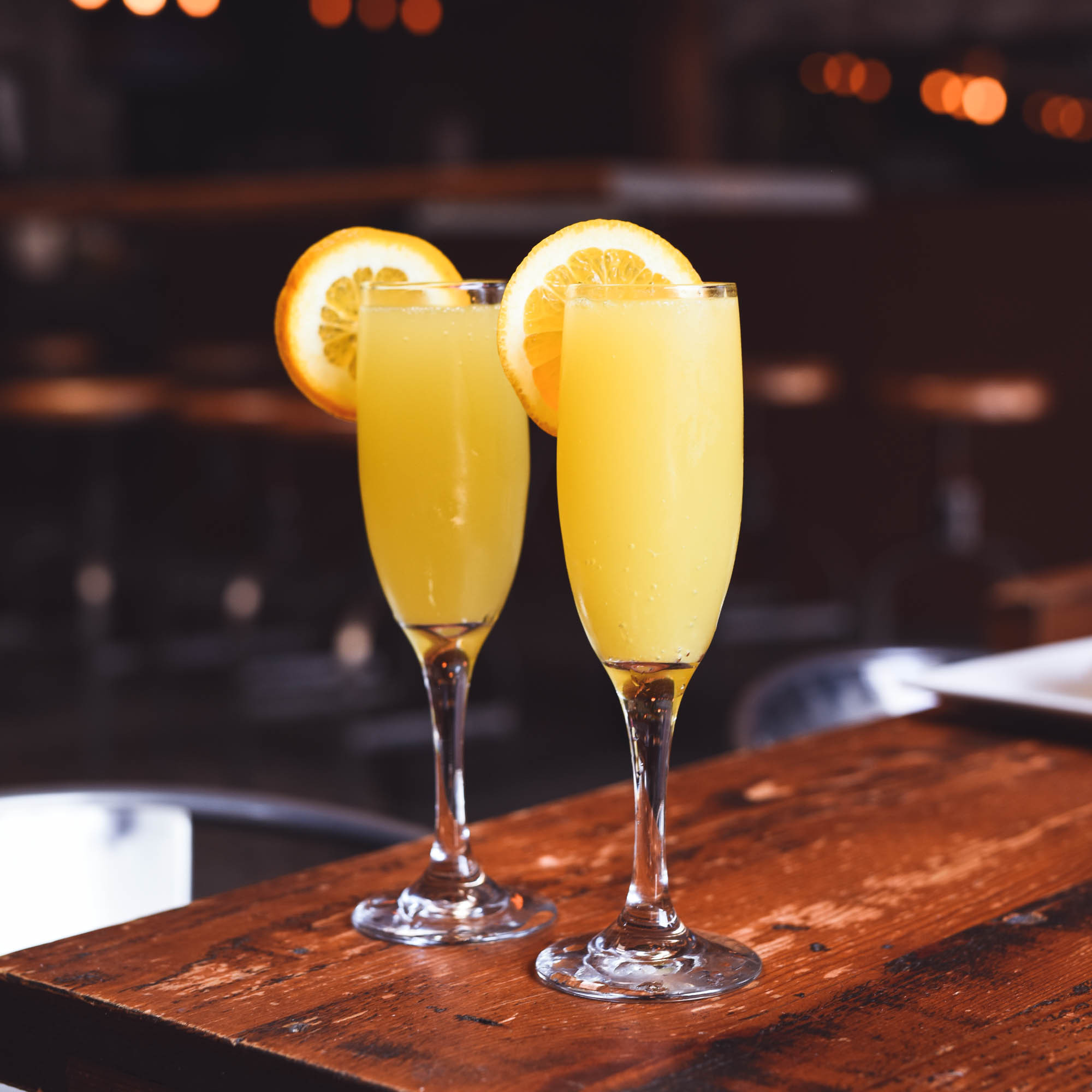 Two mimosas in champagne flutes on a wooden table with sliced orange garnish on the rim. 