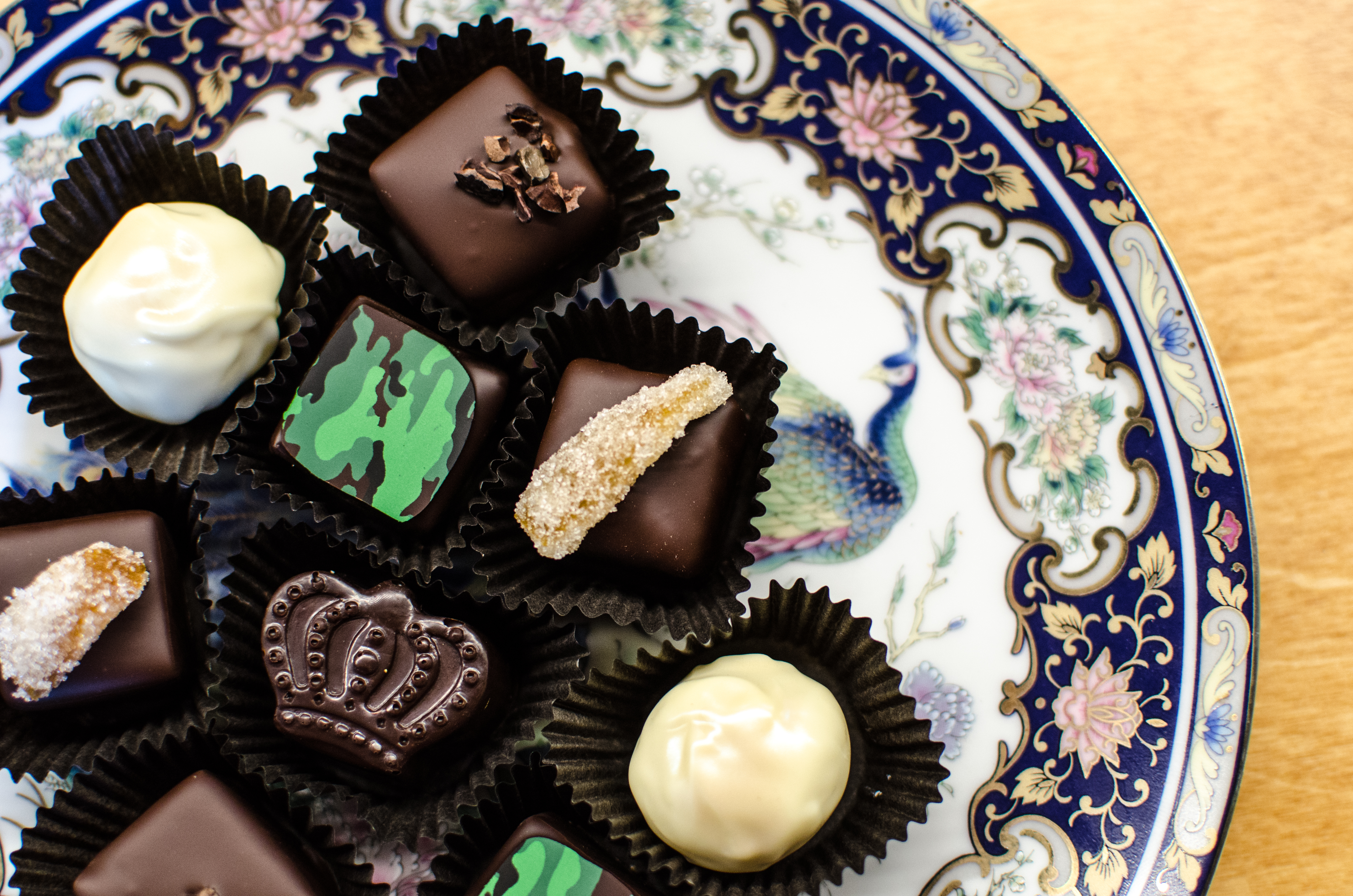Several fancy chocolates sit on an elaborate peacock plate. One chocolate is crown-shaped, several are garnished with candied ginger, and several are garnished with a camo-patterned sliver of chocolate.