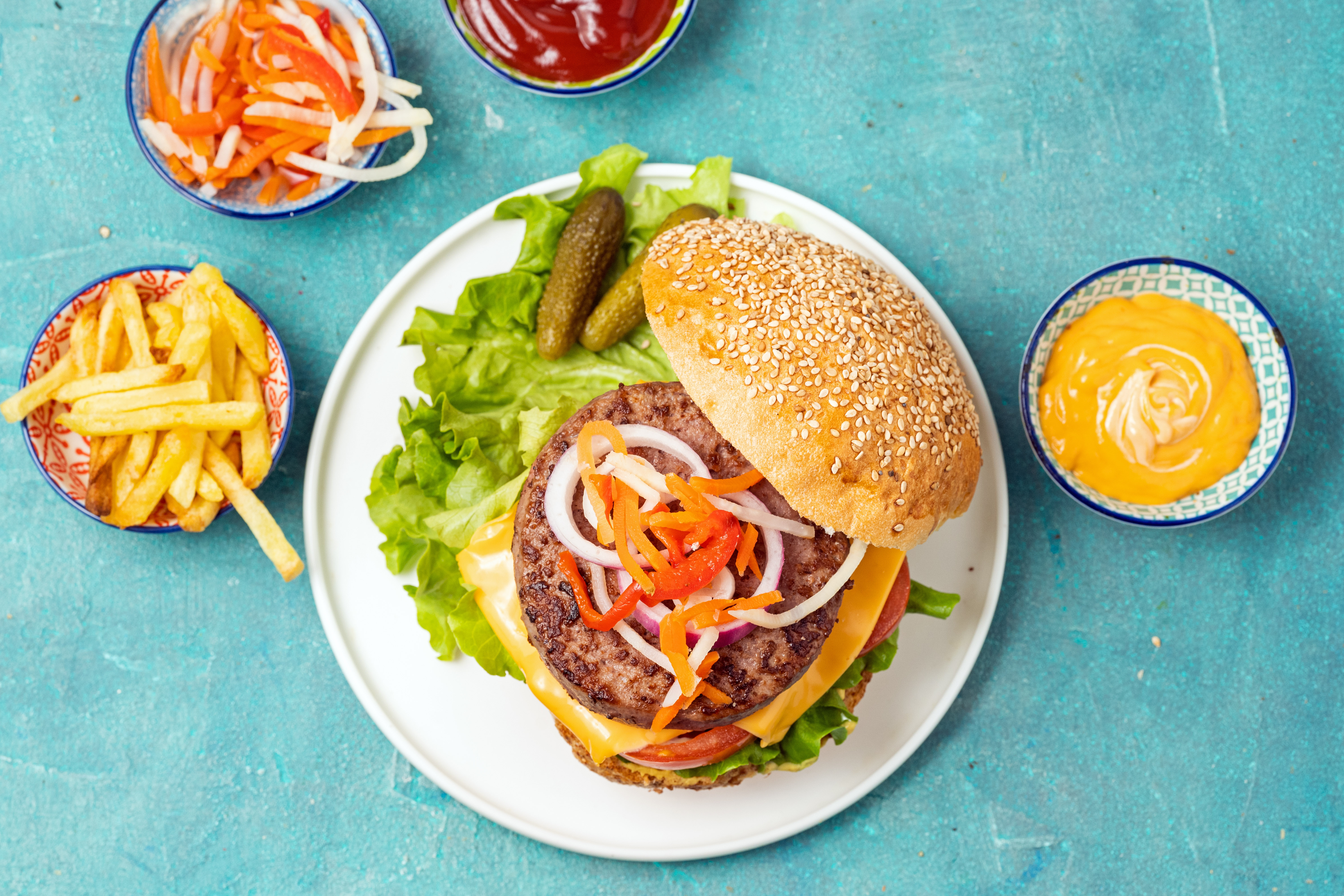 An open faced burger on a round white plate topped with onions, lettuce, cheese, and tomato with small bowls of condiments and a bowl of fries on the side with with a blue background.
