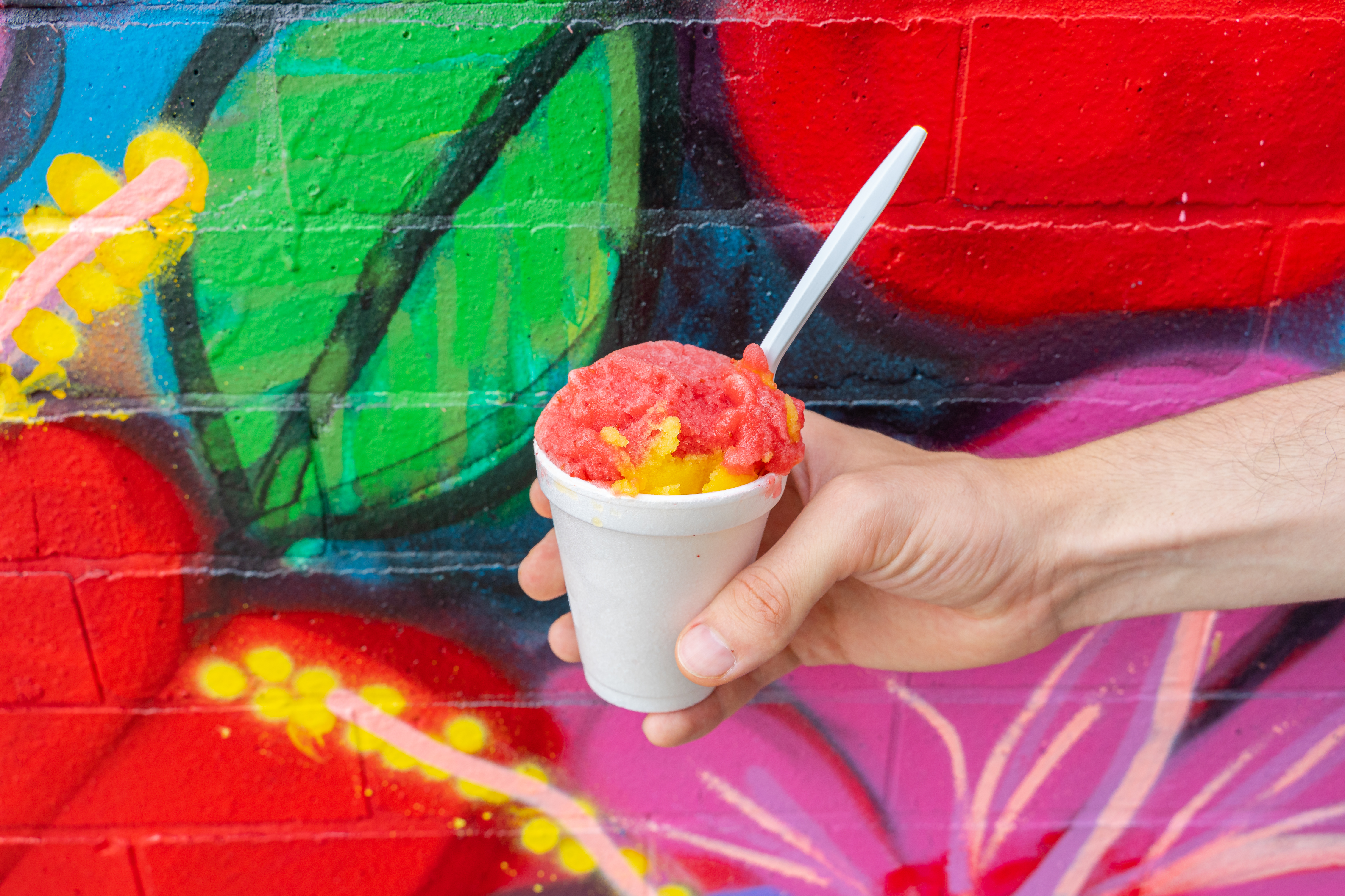 A hand holds a white styrofoam cup of red and yellow Italian ice in front of a colorful wall mural.