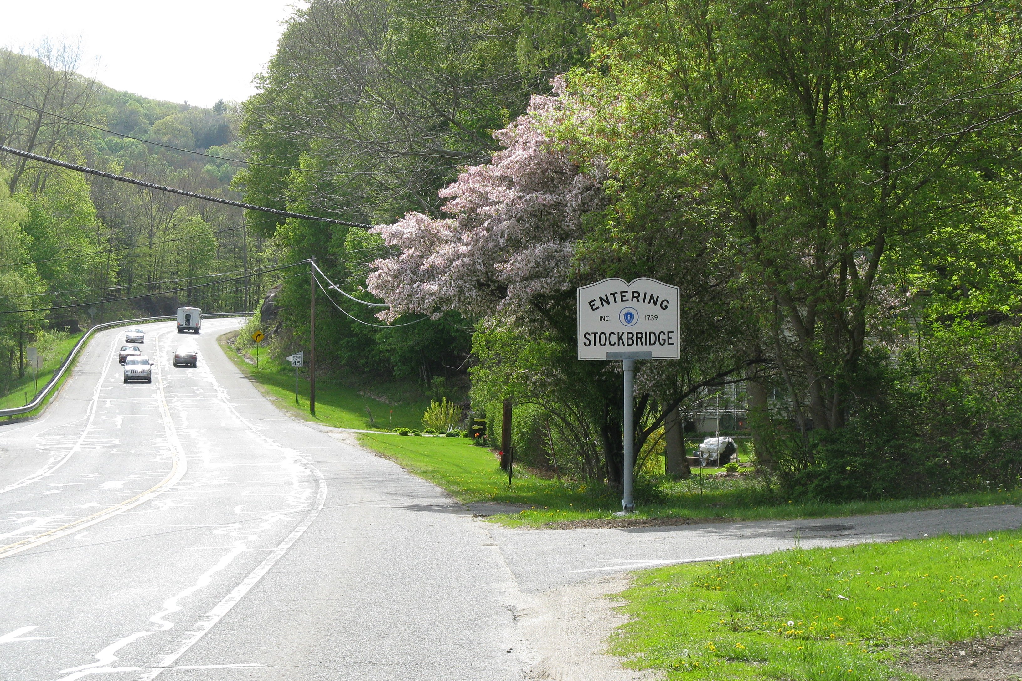 A highway with trees on both sides. Some of the trees have pink blossoms. There is a sign in the foreground that reads: Entering Stockbridge.