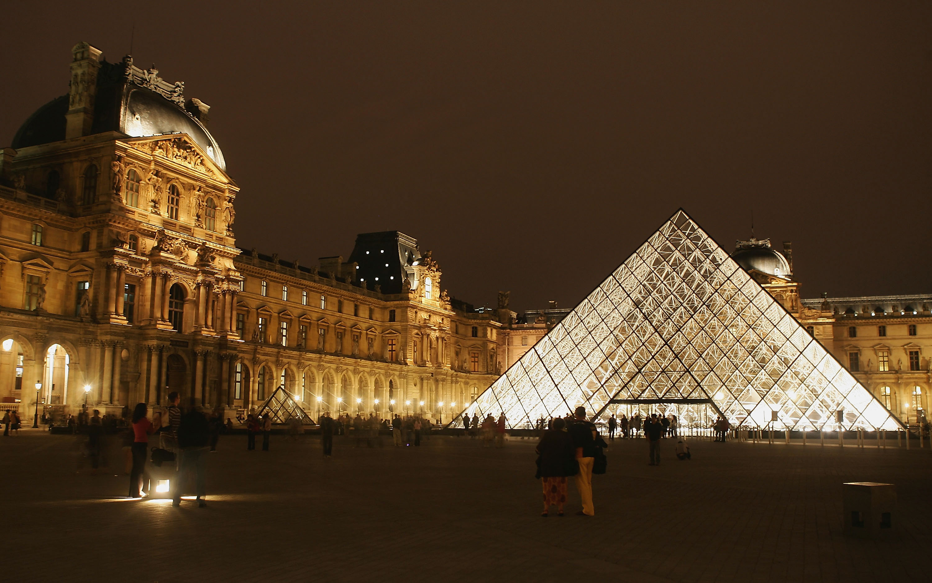 A glass and steel pyramid at night set among the courtyard of opulent French buildings. 
