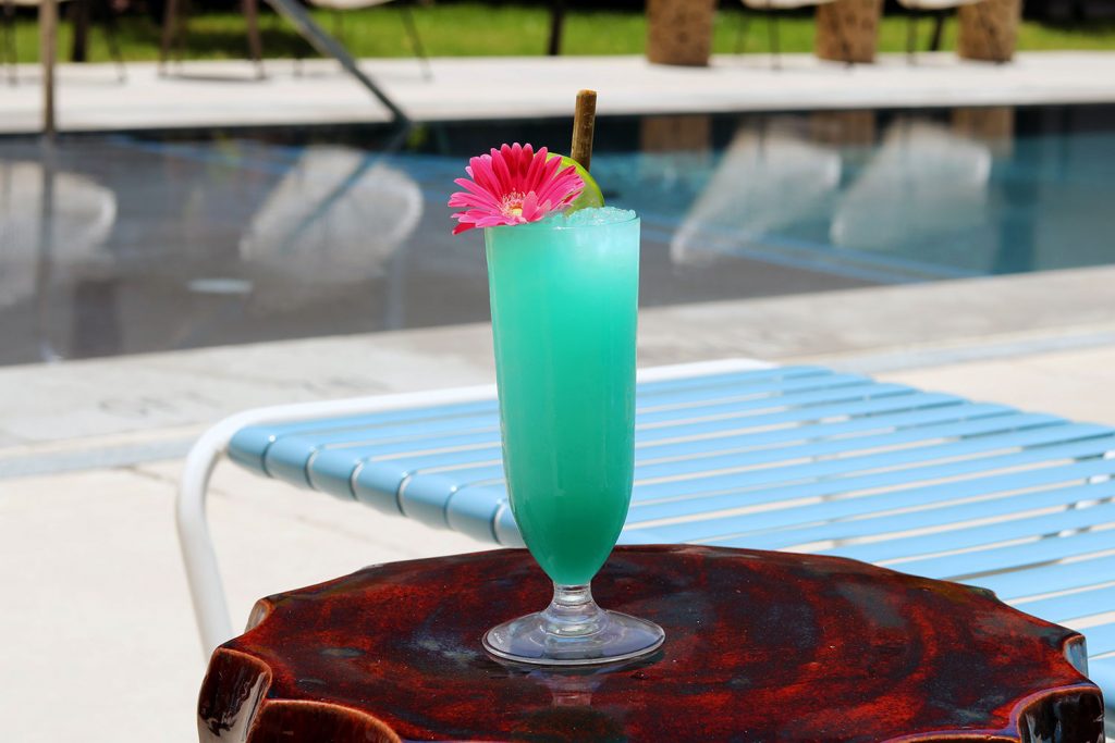 A tall blue cocktail in a glass with a pink flower in front of a swimming pool.