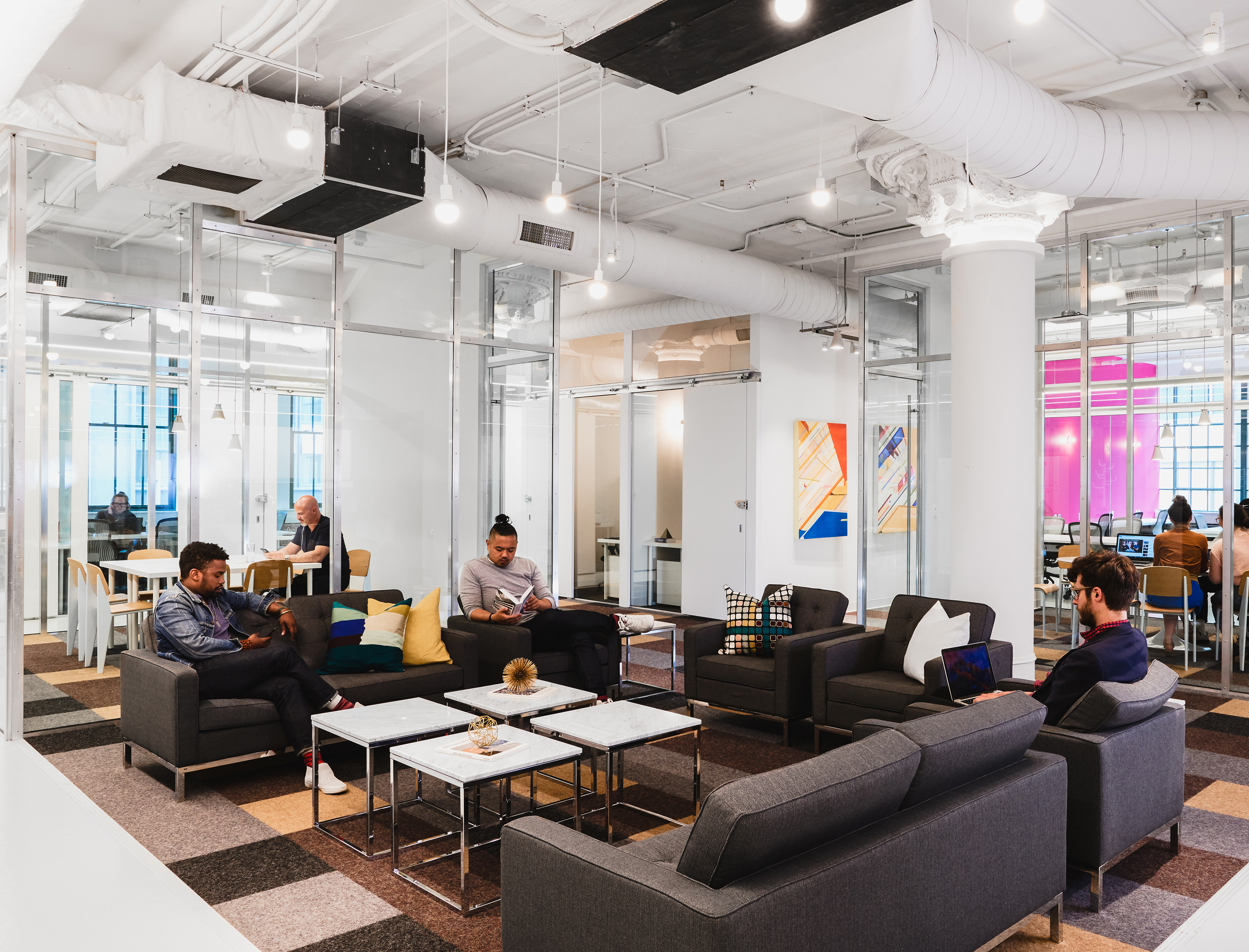 A coworking space with exposed, white ductwork and pillars, and big industrial-sized windows.