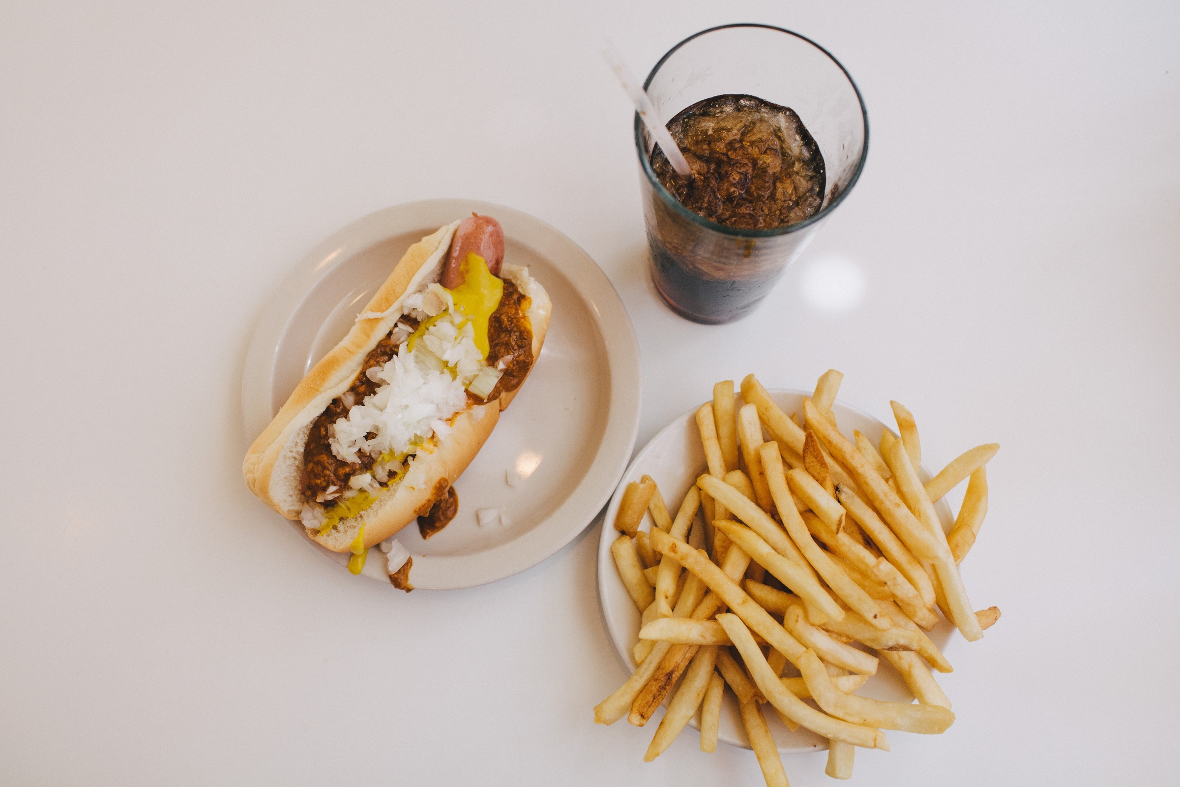 A coney dog, a plastic cup of brown cola, and a plate of french fries on a white countertop.