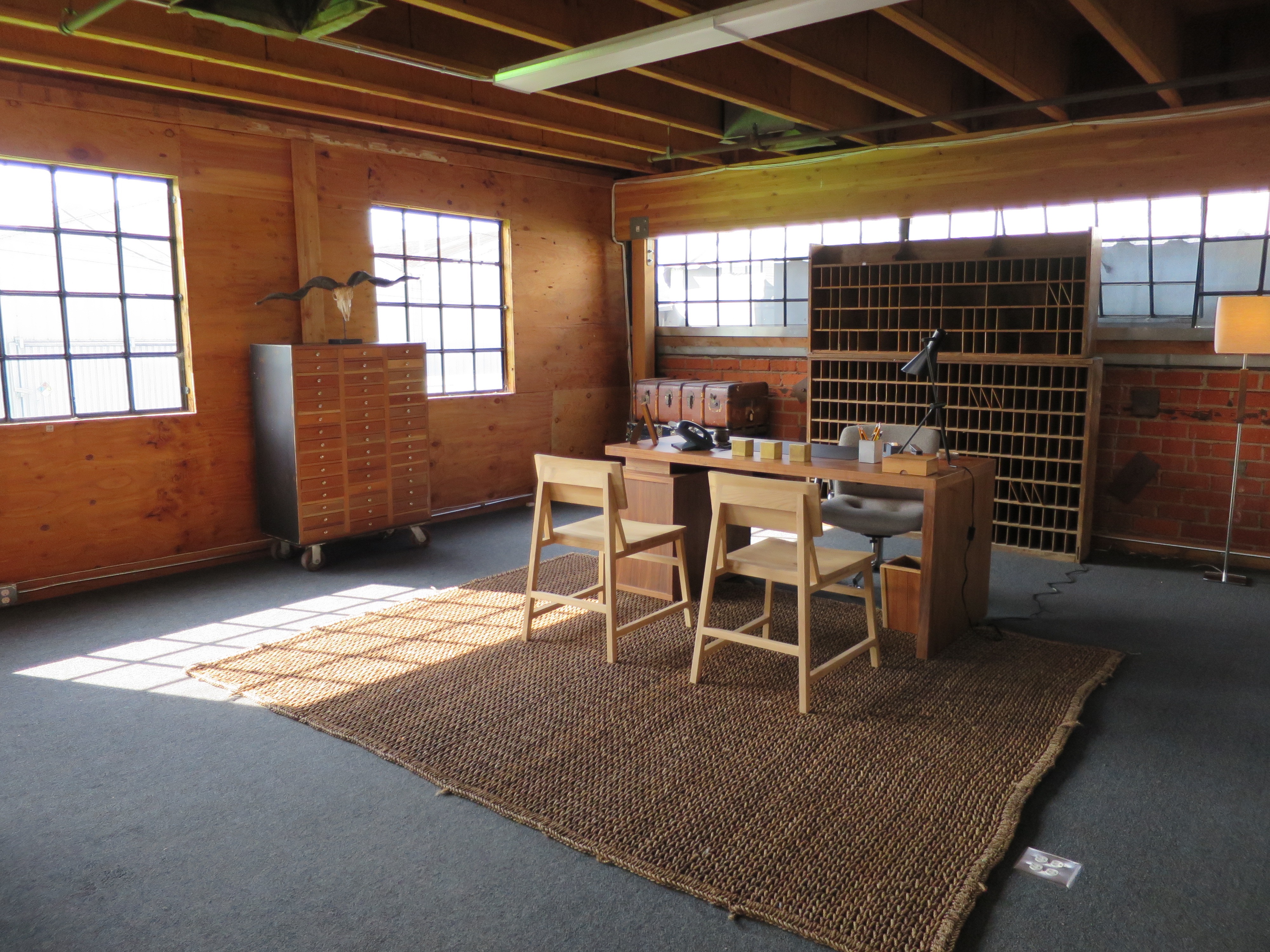 Interior of office set into corner of warehouse space with plain wood furniture.