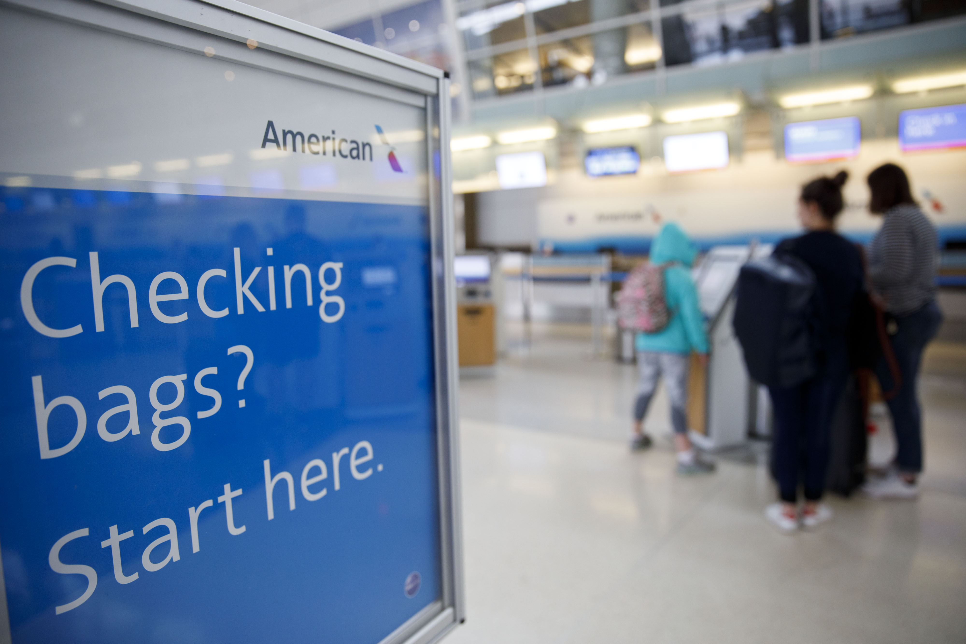 A sign for American Airlines reads: “Checking Bags? Start here.” In the background, people line up at an airport check in.