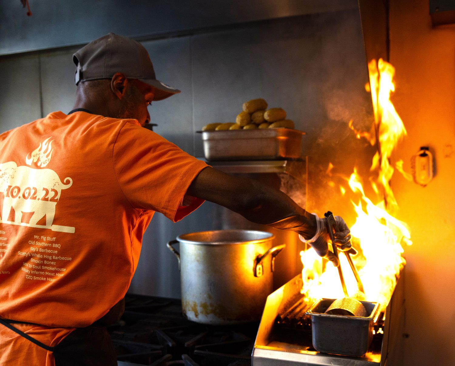 A man in an orange shirt, baseball cap and apron reaching tongs into a flaming pan.