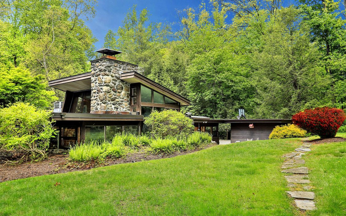 Exterior shot of angular-roofed home with large stone chimney and walls of glass set on verdant site surrounded by trees. 