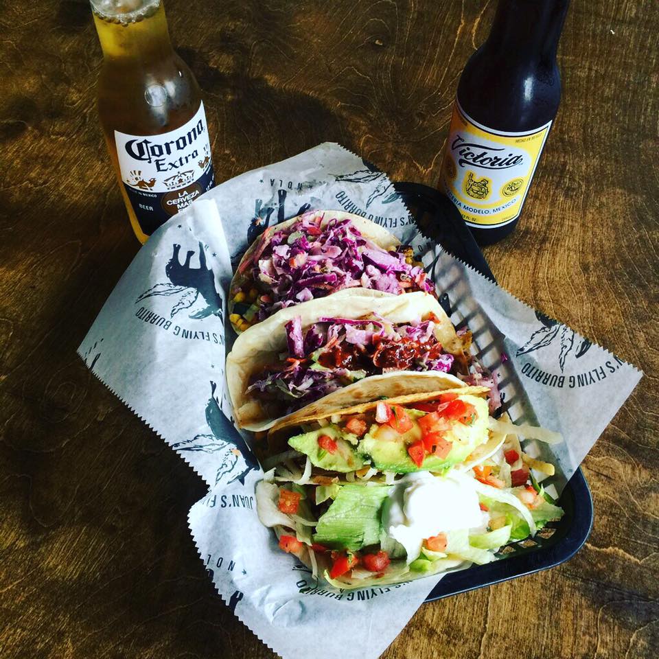 A platter of four tacos, two topped with red cabbage and two with tomatoes and iceberg lettuce, on a table with two bottles of beer.