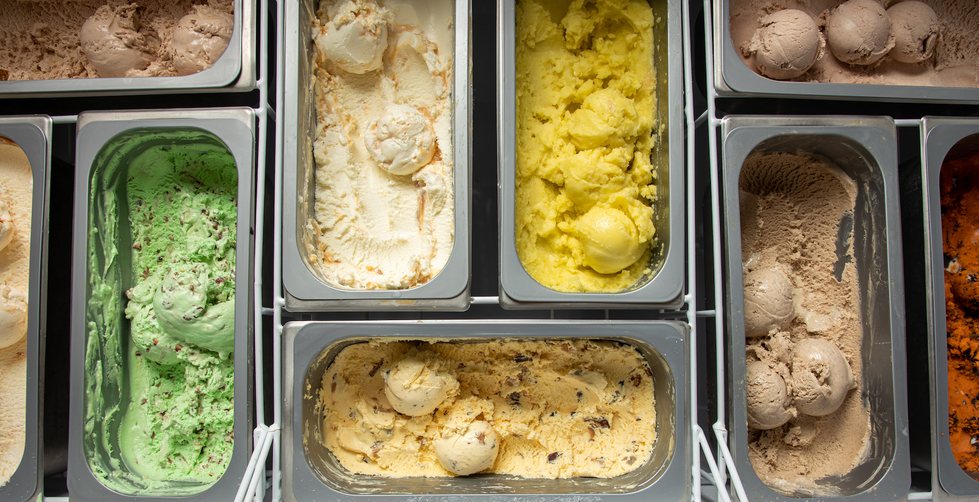 A case of seven different ice cream flavors lined up in metal bins, in green, yellow, and cream colors. 