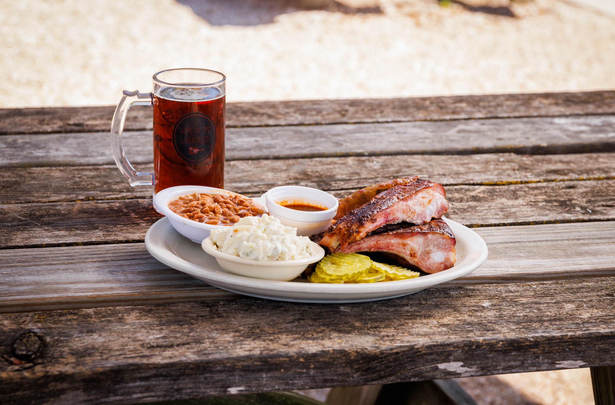 An old wooden picnic table holds a plate of ribs, beans, potato salad, and a red beer.