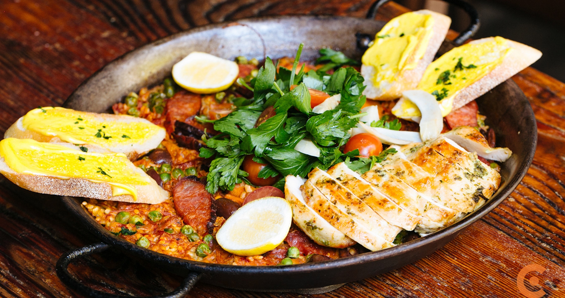 A metal paella dish filled with rice and topped with chicken, chorizo, tomatoes, greens, lemon wedges, and slices of bread.