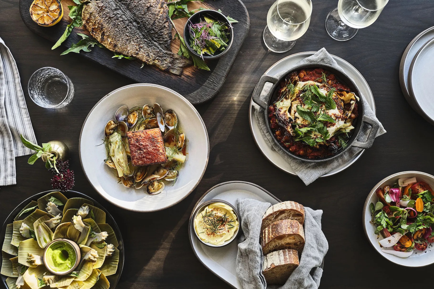 A top-down shot of plates of wood-fired food, bread, and a whole fish.