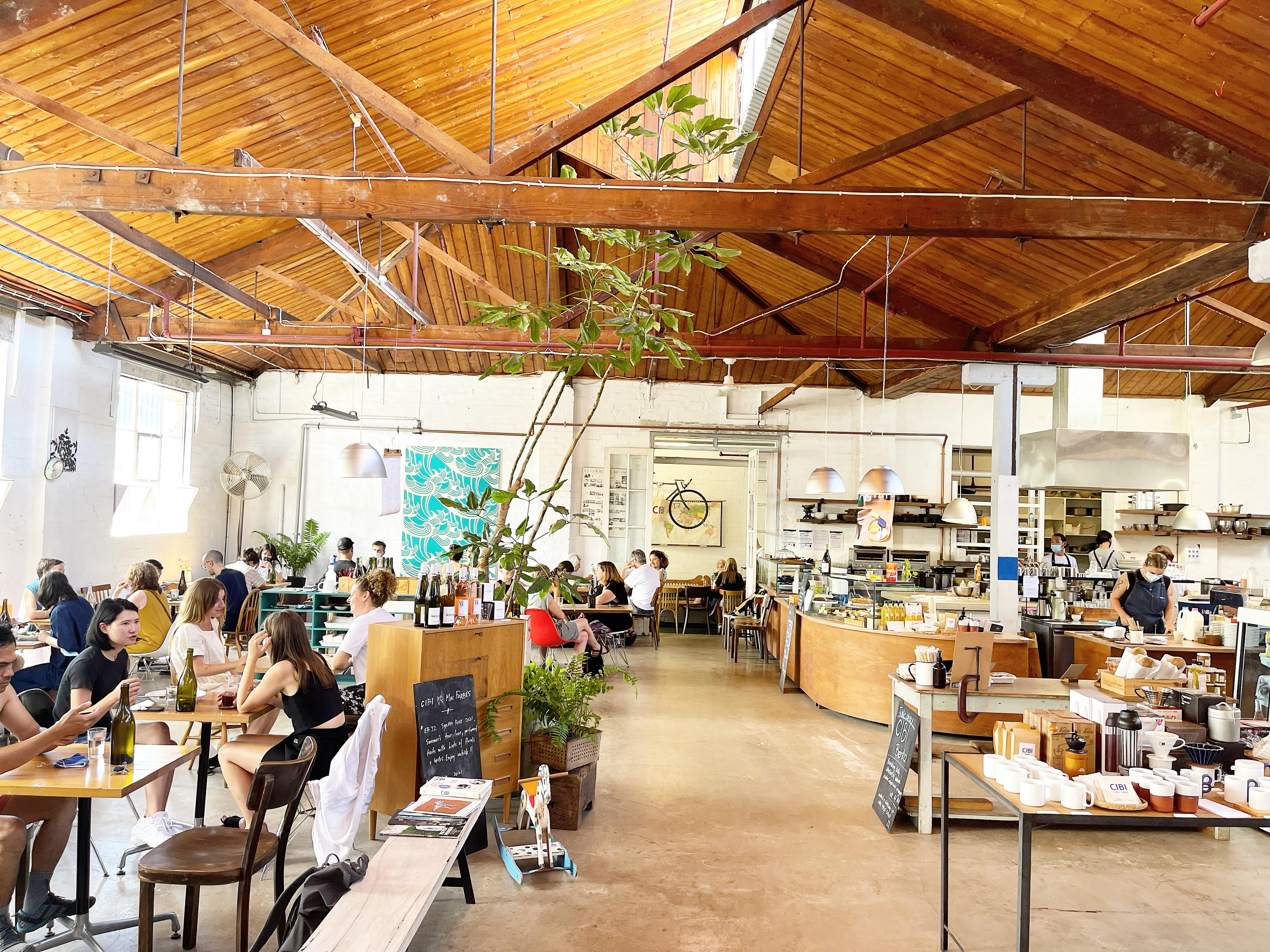 A tall airy cafe, where diners sit at tables, large plants hover, and items for sale are on display.