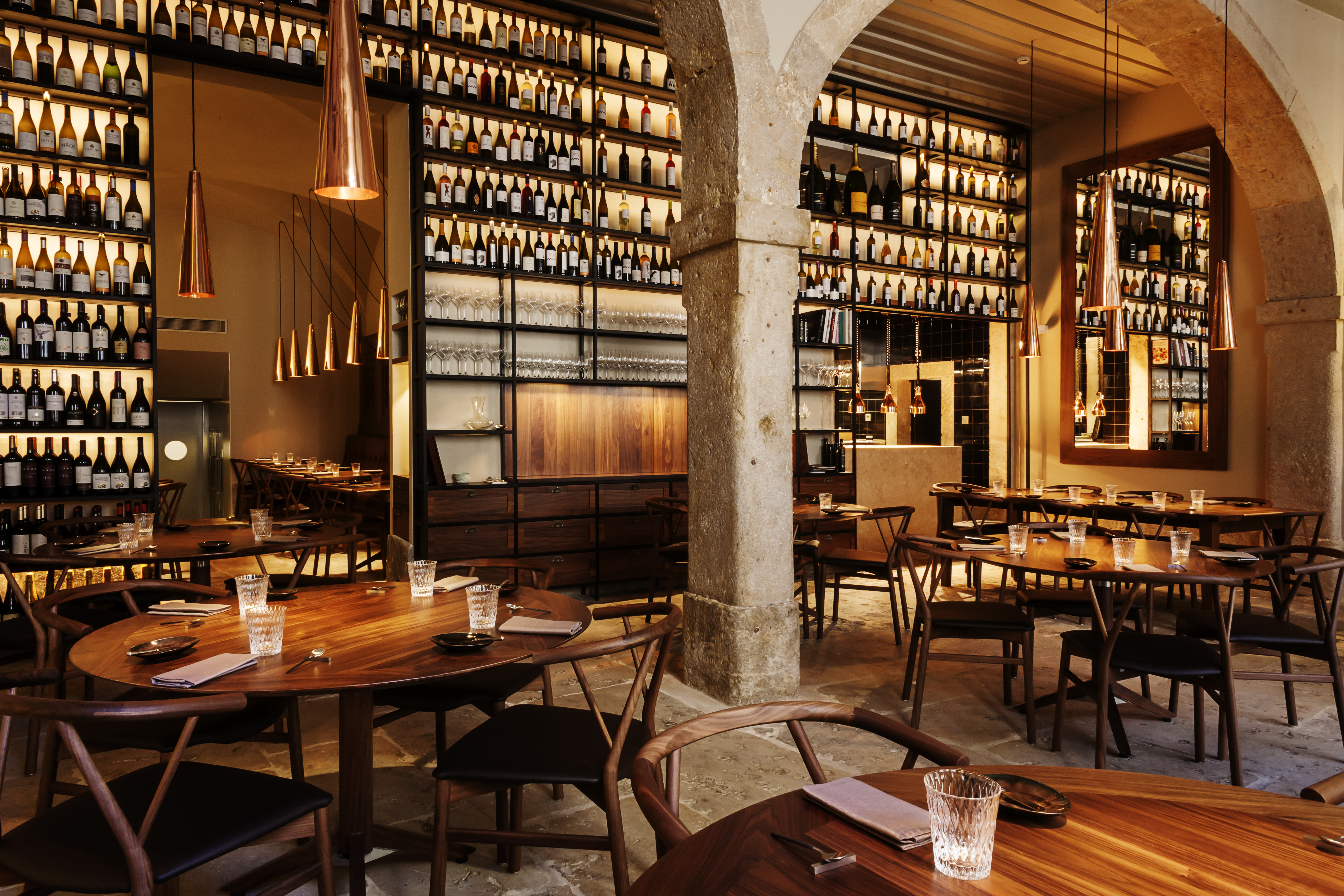 A tall restaurant interior with shelves of bottles, old stone columns, and wooden tables set for dinner. 