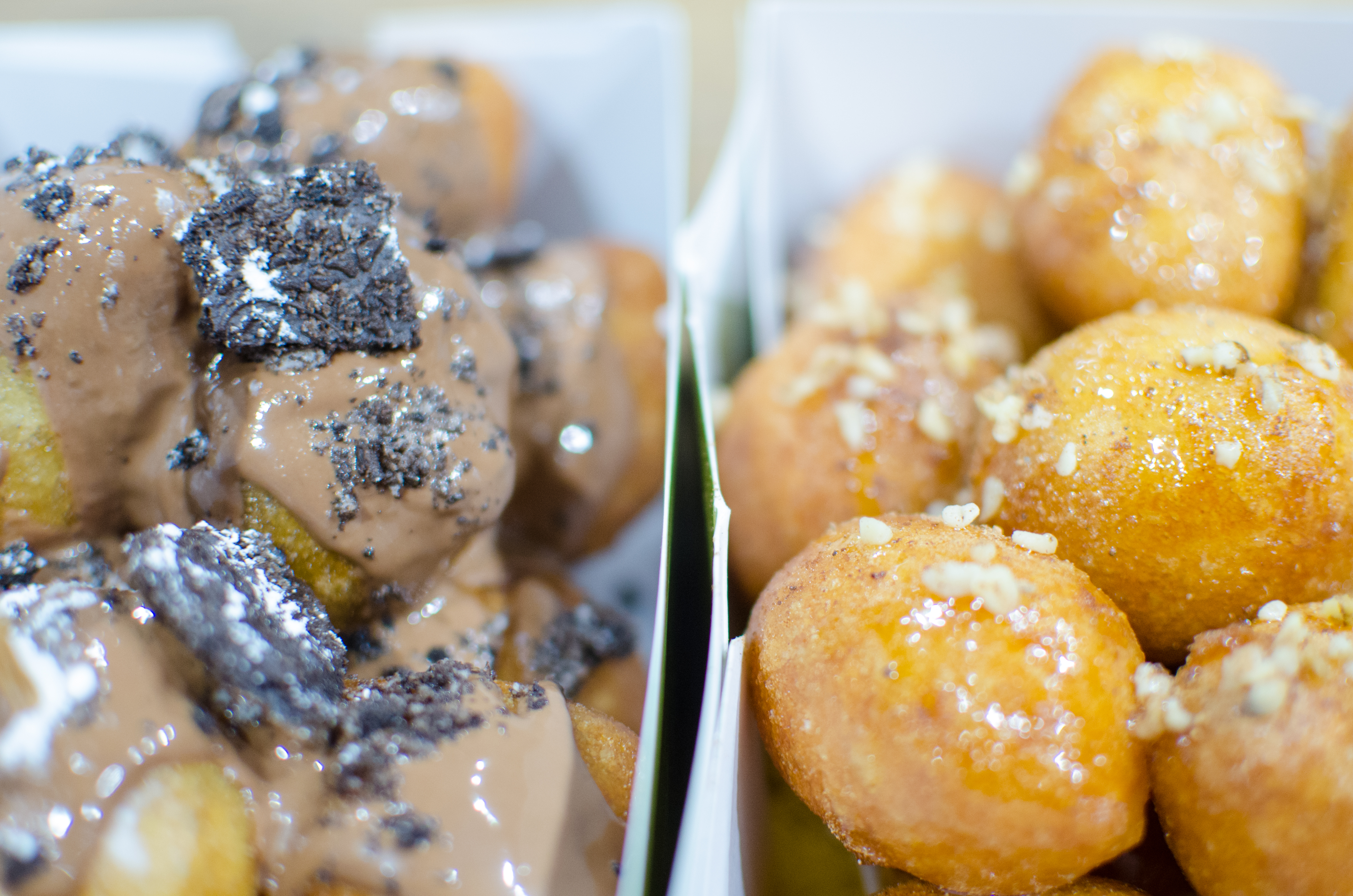 Closeup shot of two side-by-side paper containers of fried dough balls. One is topped in a chocolate glaze with crushed Oreos.