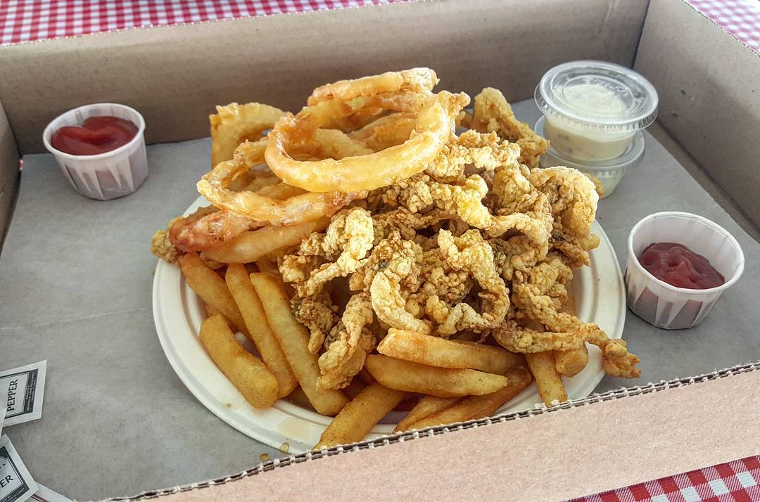 A paper plate of fried clams, fries, and onion rings sits in a cardboard tray on a red-and-white-checkered tablecloth