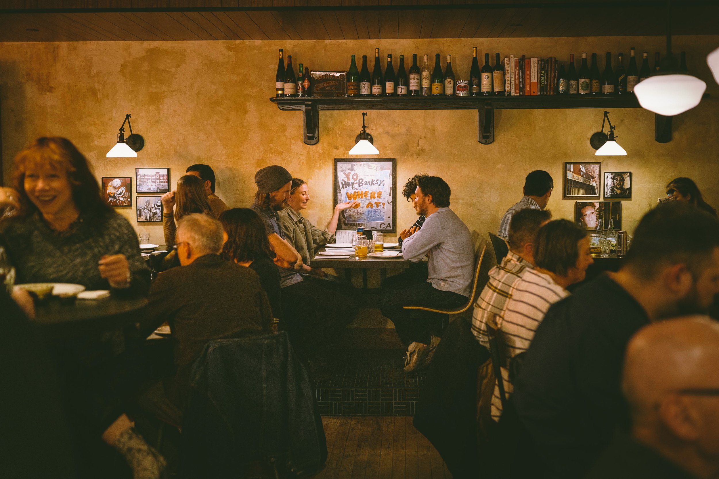 People dine at small tables inside a dimly lit restaurant with a shelf of bottles overhead