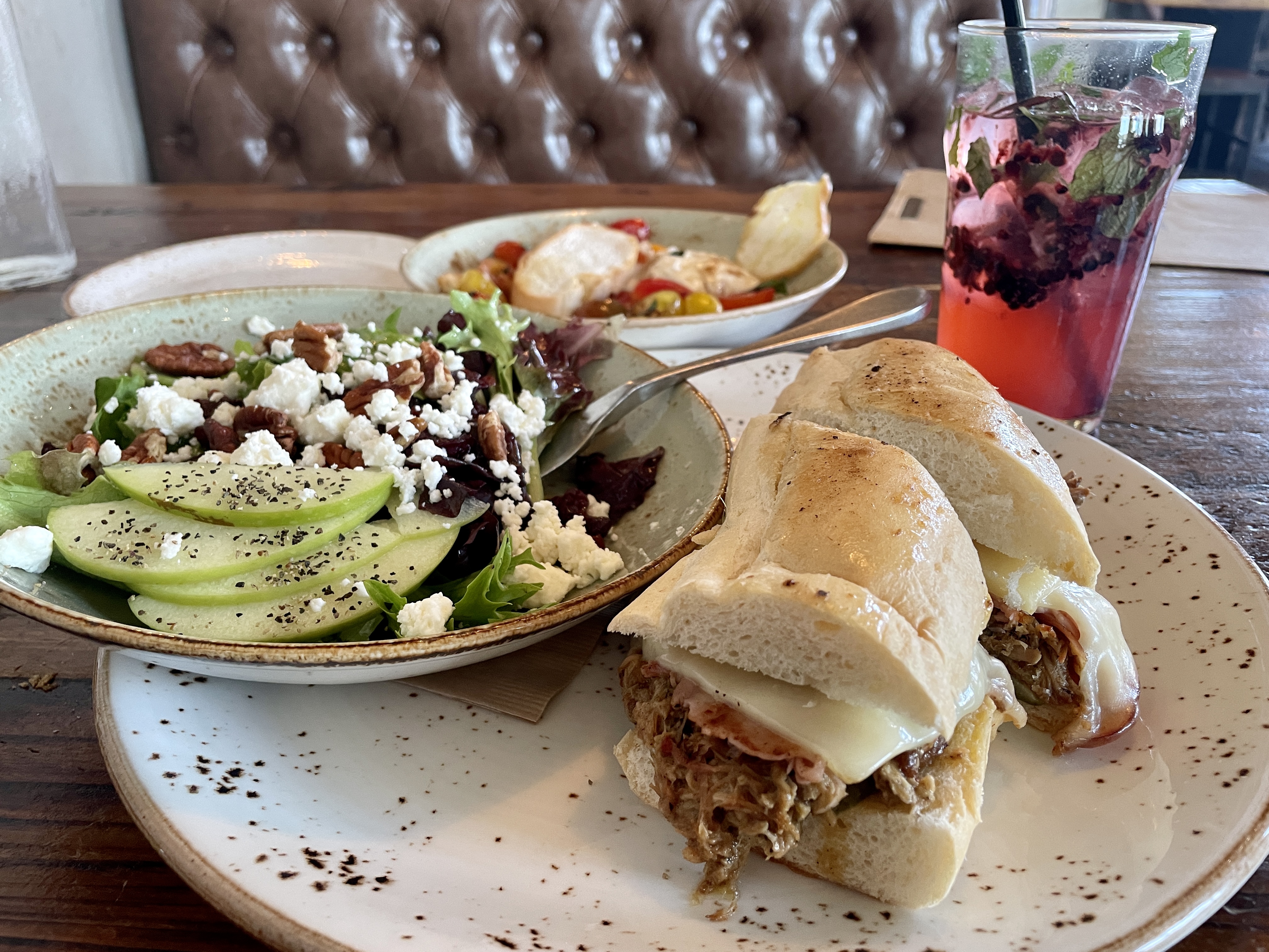A plate holds a cuban sandwich with a small dish holding a salad next it it, which is covered with green apples, crumbled goat cheese, and dried cranberries. In the background are a water glass with a drink that holds smashed blackberries and a small plate of burrata, tomatoes, and bread.