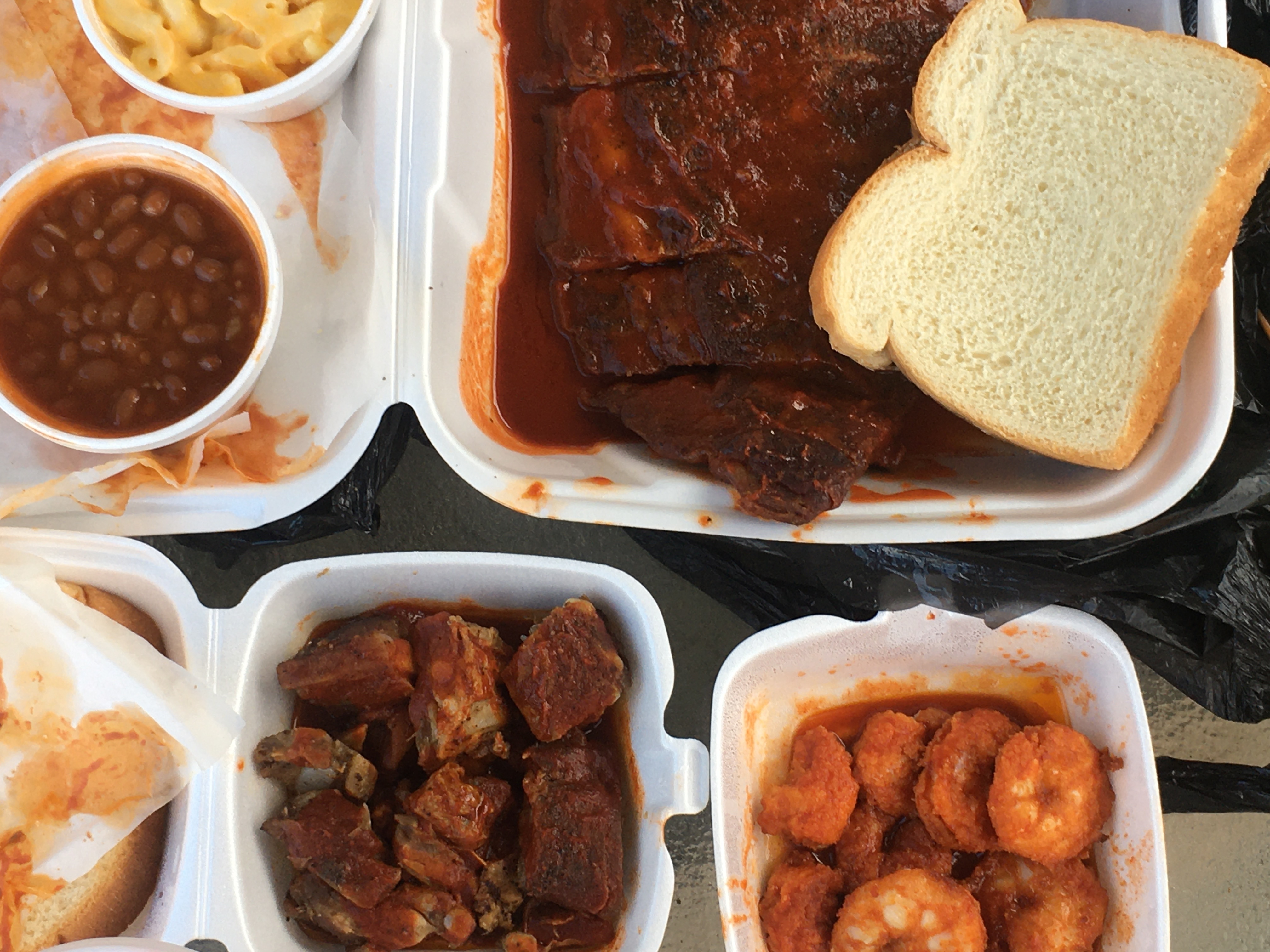 Ribs with white bread, rib tips, and fried shrimp in pools of red sauce in takeout containers.