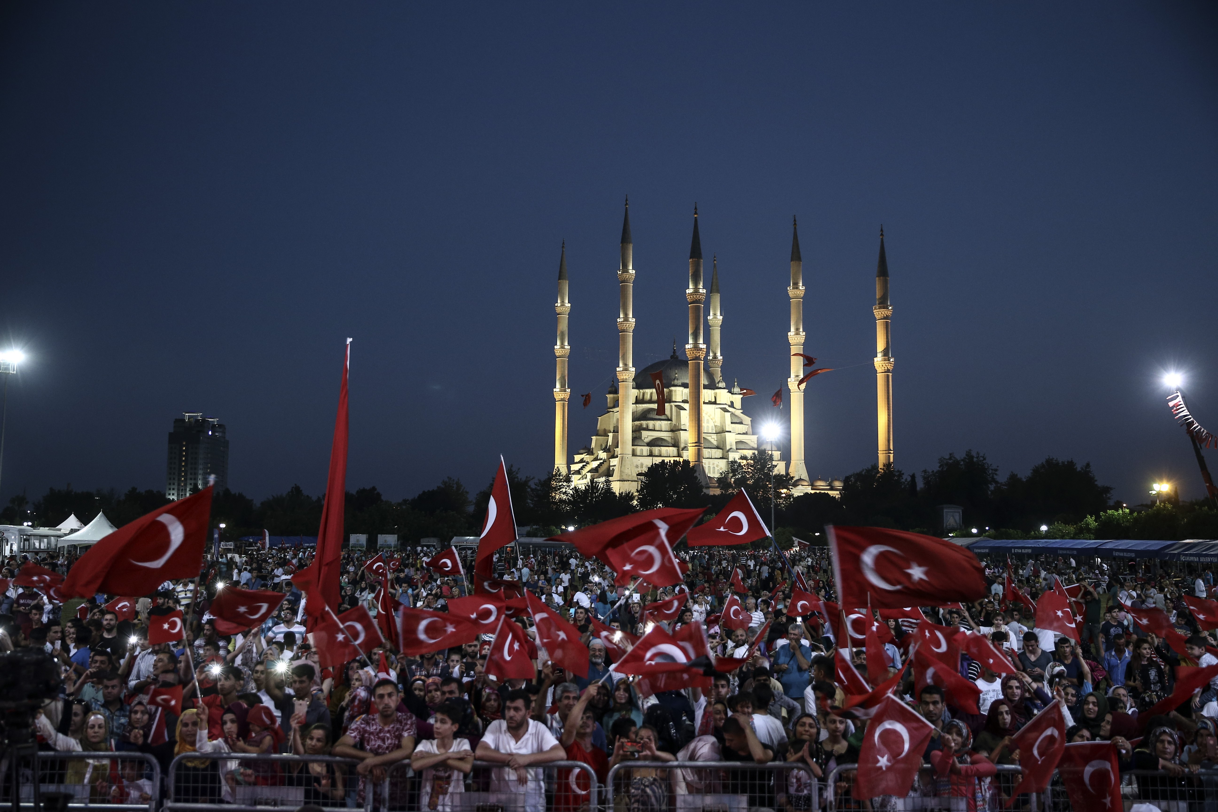 Demonstrators hold up Turkish flags at dusk and an illuminated mosque stands in the background. 