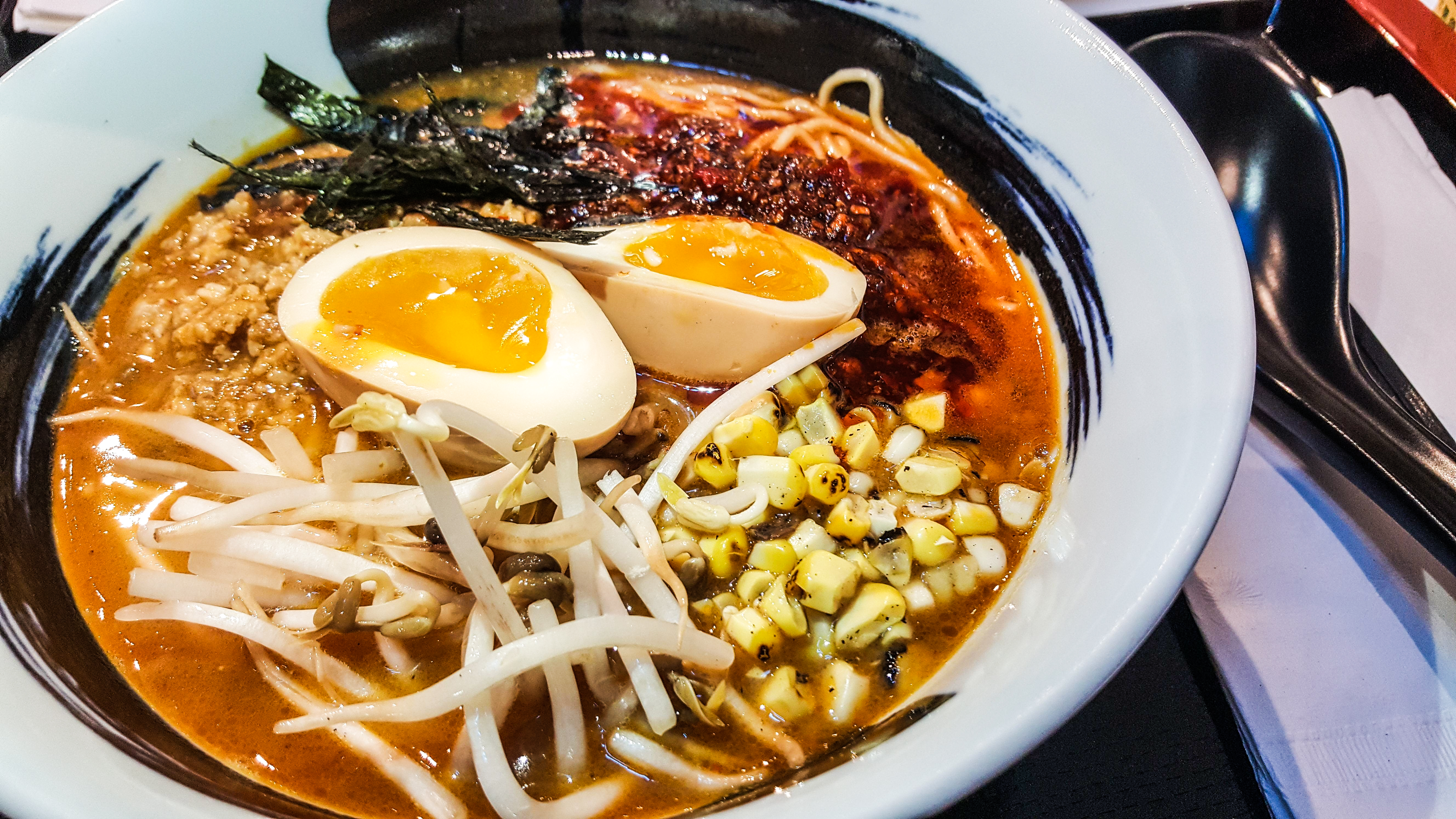A white bowl with a thick, ink-like swoosh of black around the rim is filled with spicy miso ramen, including corn, bean sprouts, and egg, and other ingredients.