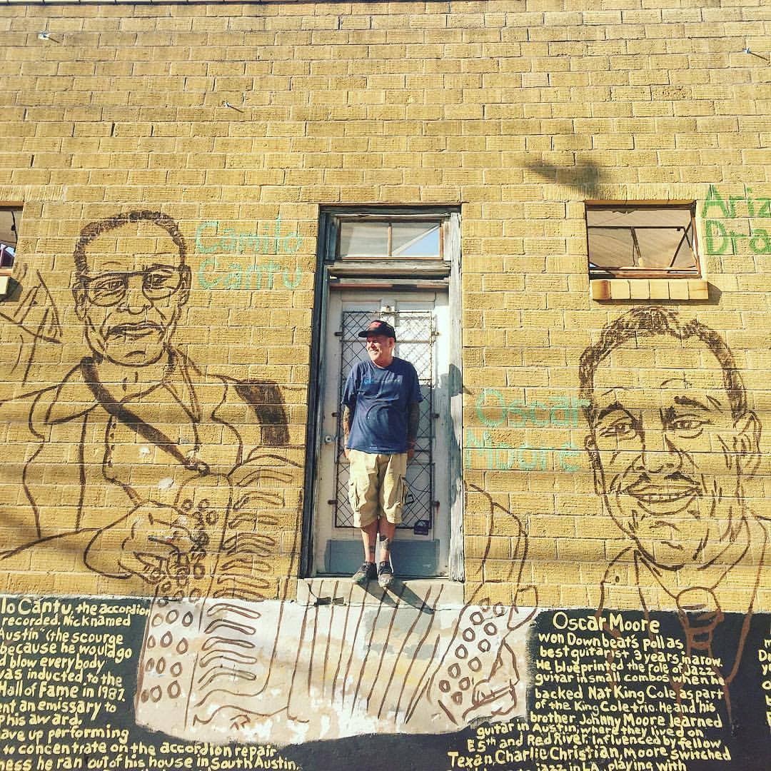 A man in ball cap, T-shirt and jeans in front of a yellow brick wall with a mural  of two men