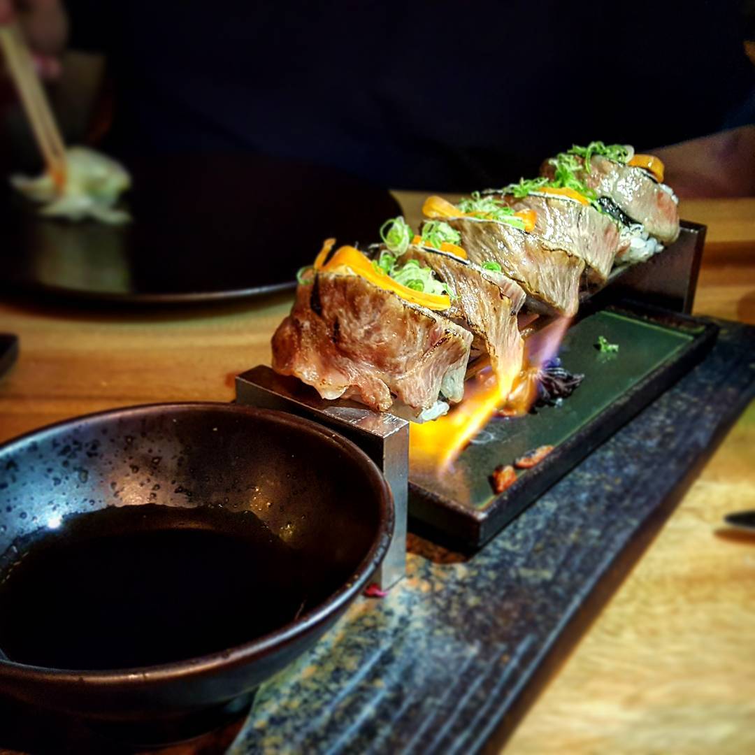 Slices of wagyu beef above a flame with a small dark bowl filled with dipping sauce