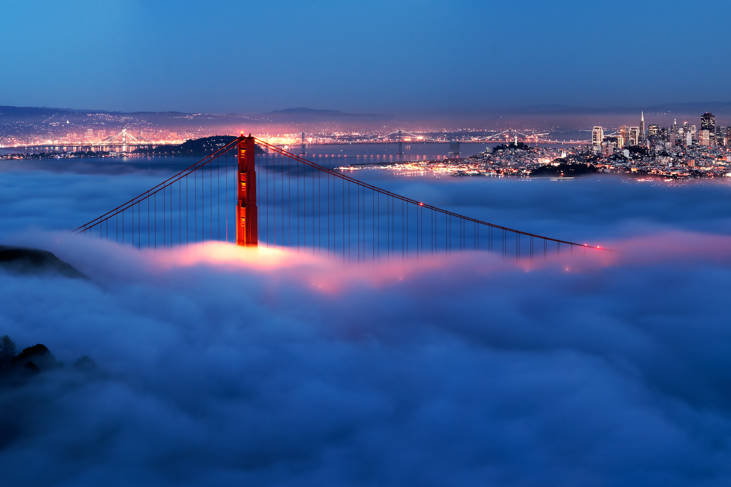A tower of the Golden Gate Bridge peaking above the fog with the lit-up skyline of SF and Oakland behind.