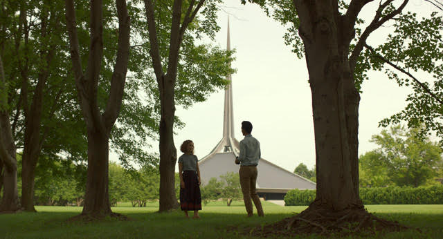 Photo of a man and woman standing amid trees in front of large steepled structure.