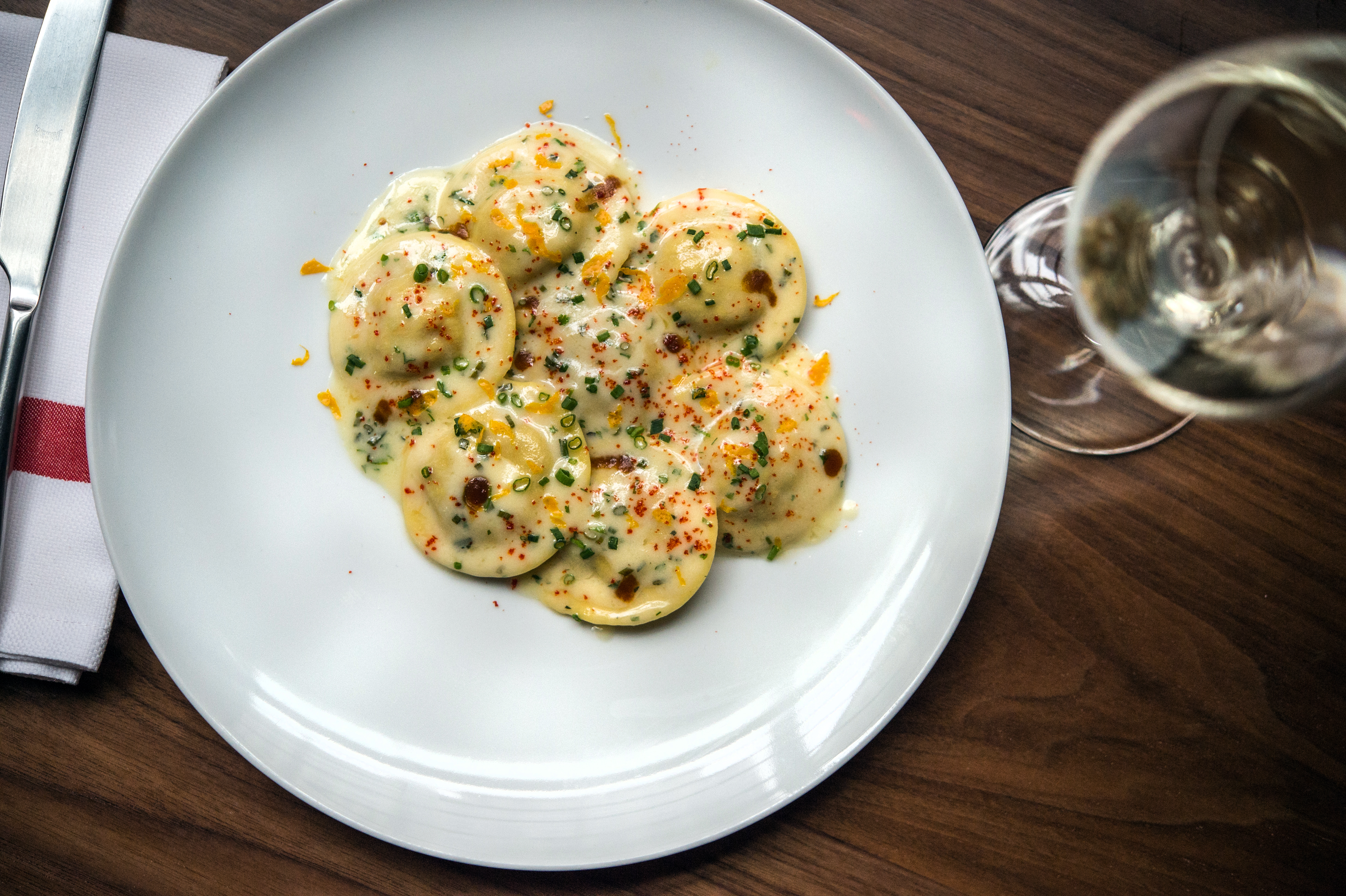 A saucy pile of lobster ravioli on a white plate with a wine glass to the right.