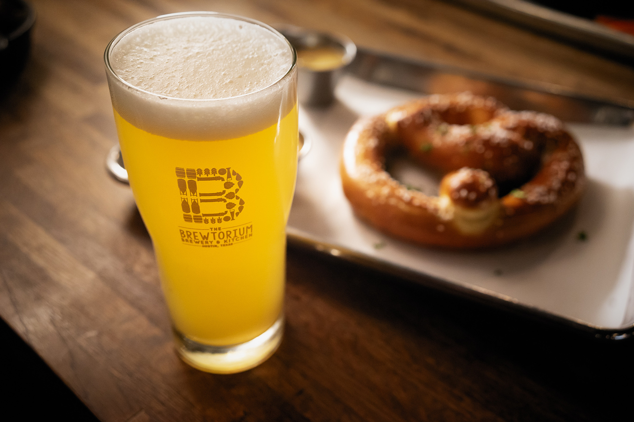 A glass of cloudy yellow beer in a glass with the words Brewtorium in front of a blurry pretzel.