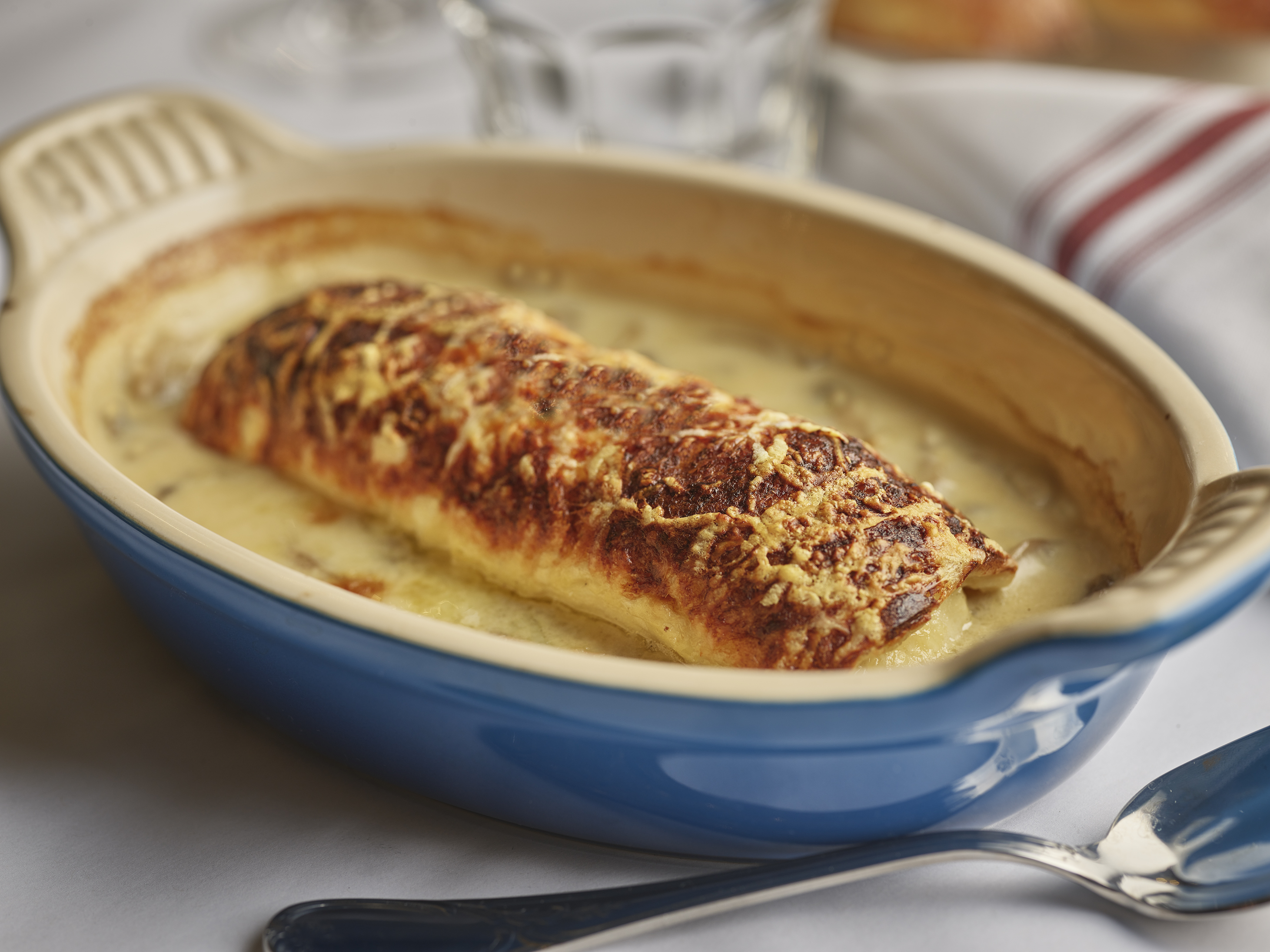 A blue oval dish filled with one long quenelle with a burnt top, sitting in a creamy white sauce.