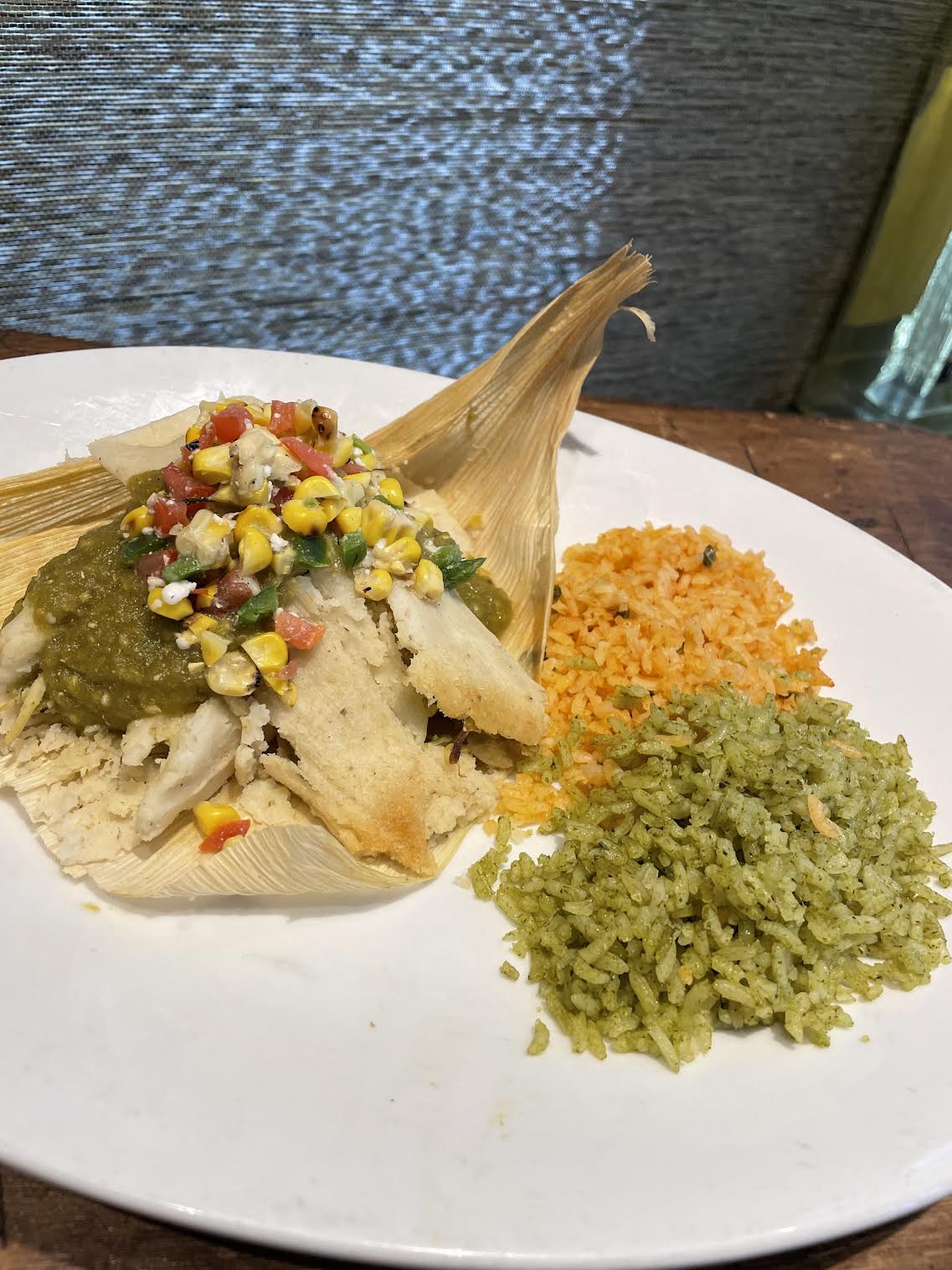 A tamale is on a plate and peeled, with some of it forked into. Its topped with corn and pico de gallo. Next to it on the plate is green rice and orange rice.
