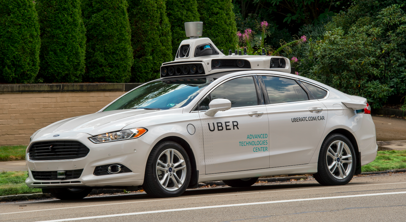 An Uber self-driving model car is shown parked by a tree-lined sidewalk. 