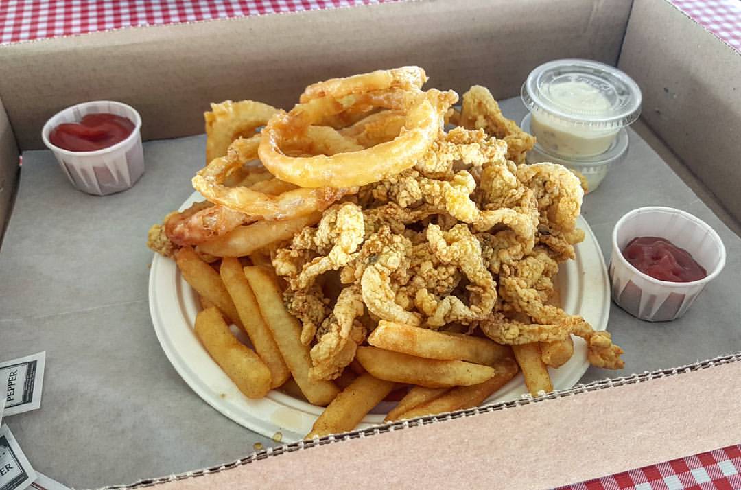 On top of a red and white checkered tablecloth, a cardboard tray is stacked high with fried onion rings, clams, and fries, with little paper cups of red ketchup to the side.